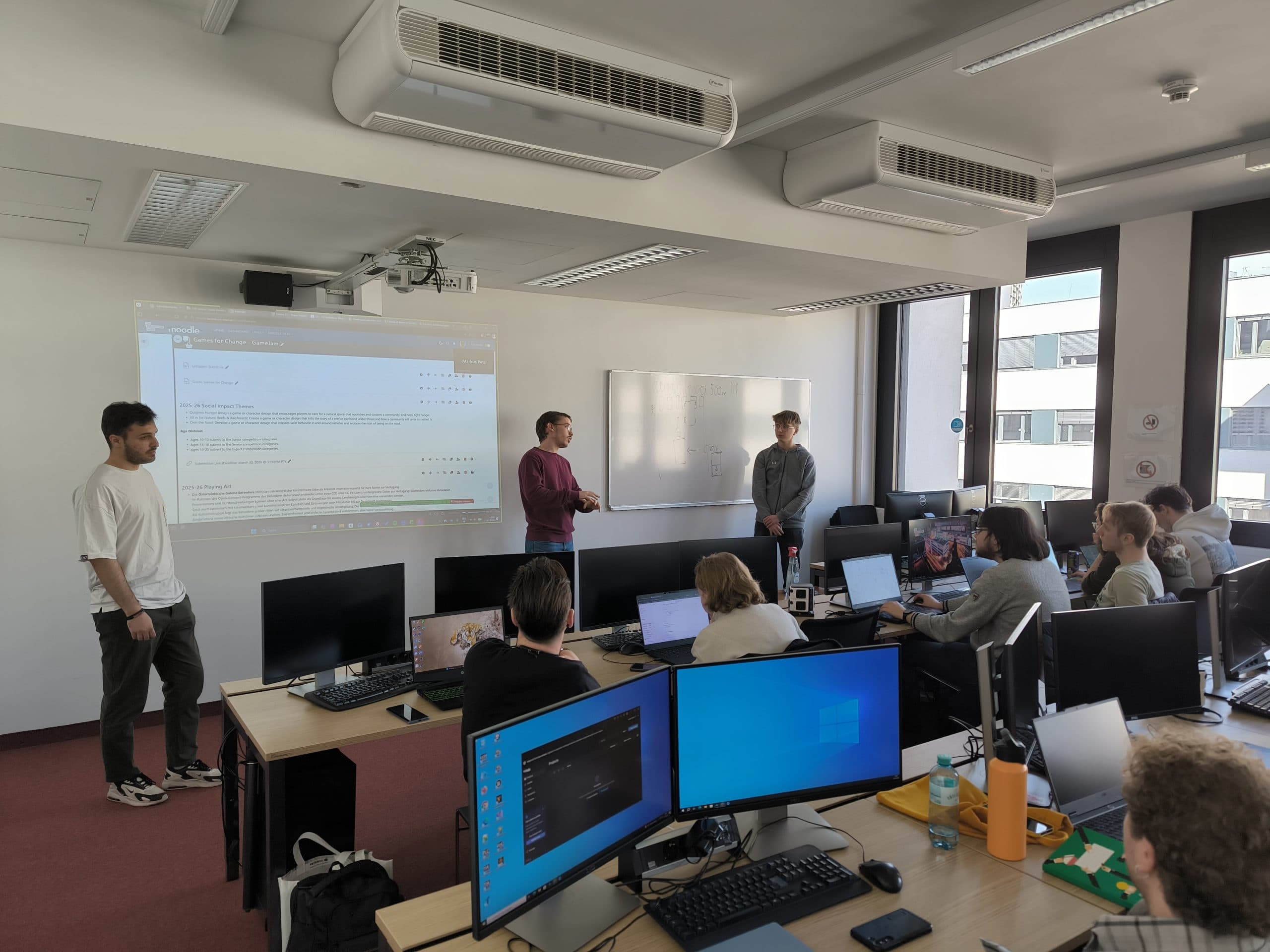 Ein Klassenzimmer mit Schülern an Computern, die drei Personen gegenüberstehen, die an der Stirnseite präsentieren. Ein Projektor zeigt Inhalte auf einer Leinwand an, und durch große Fenster auf der rechten Seite fällt Sonnenlicht ein.