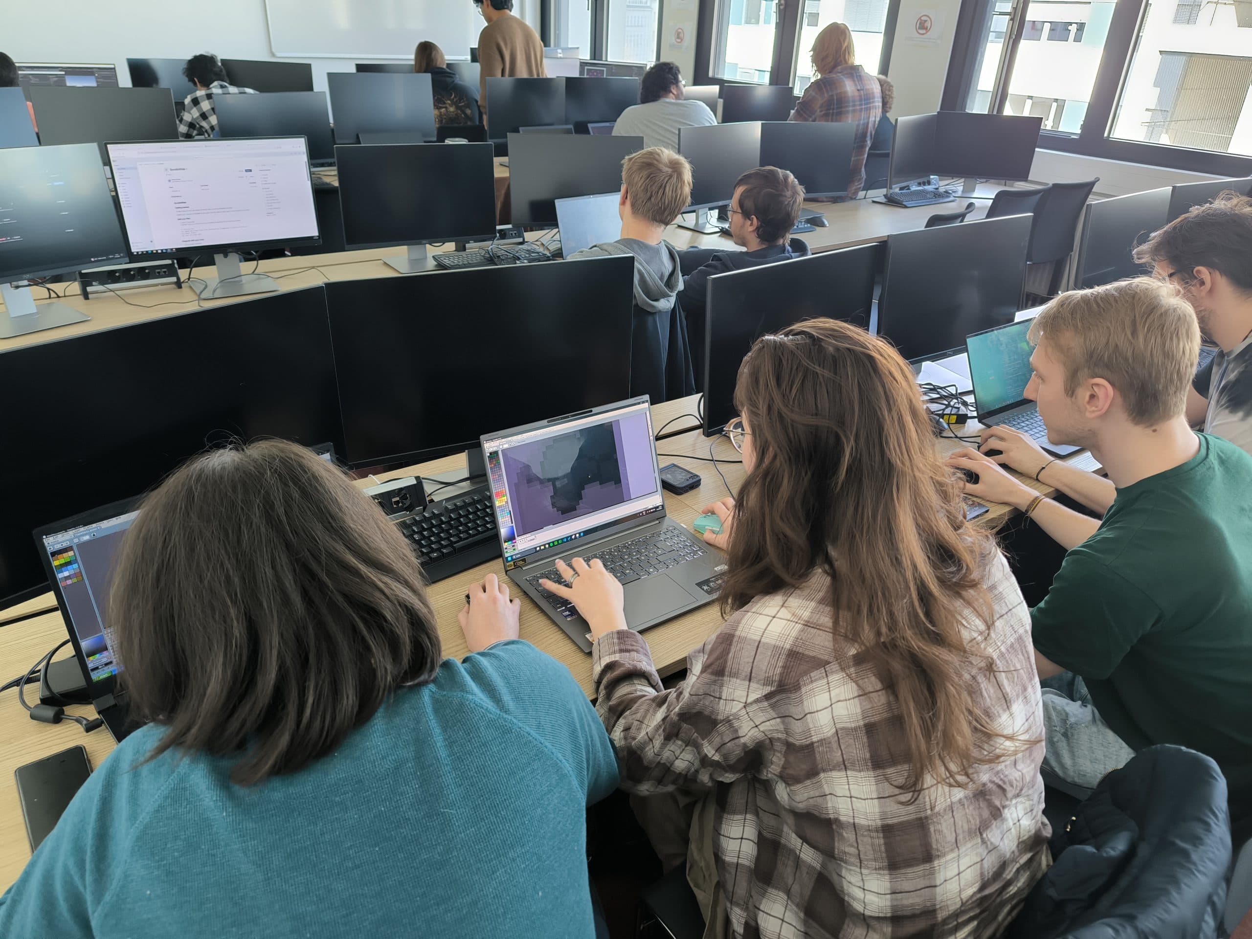 Studierende sitzen an Tischen mit Laptops und Desktop-Monitoren in einem Computerraum. Einige arbeiten an 3D-Grafik- oder Designsoftware, andere konzentrieren sich auf ihre Bildschirme. Der Raum ist hell und mit natürlichem Licht gefüllt.