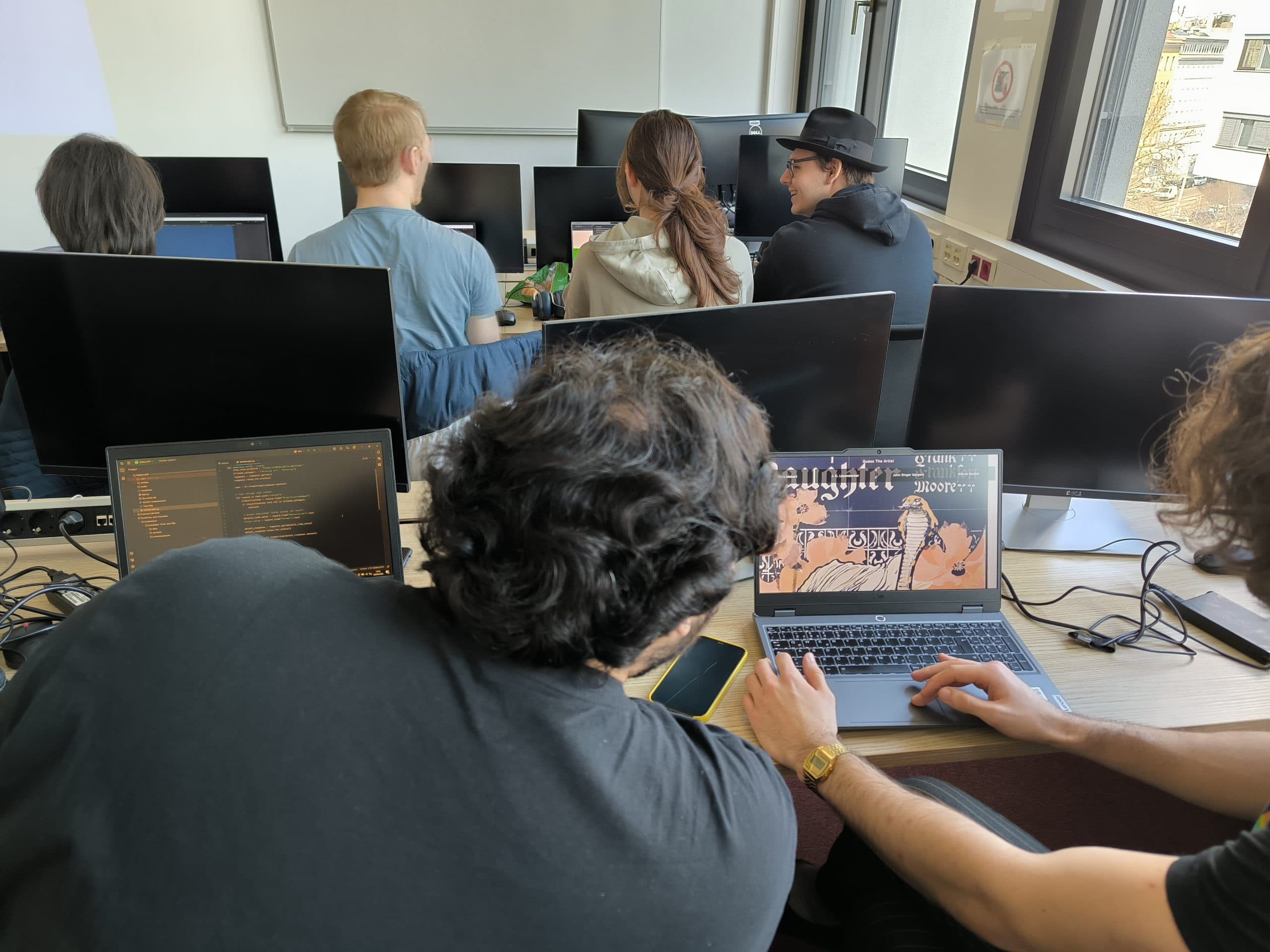 Mehrere Personen sitzen an Tischen mit Computermonitoren in einem Klassenzimmer. Zwei Personen im Vordergrund schauen auf einen Laptop, auf dem ein Code und eine Grafik angezeigt werden, während andere nach vorne blicken und sich untereinander unterhalten. Sonnenlicht strömt durch das Fenster.