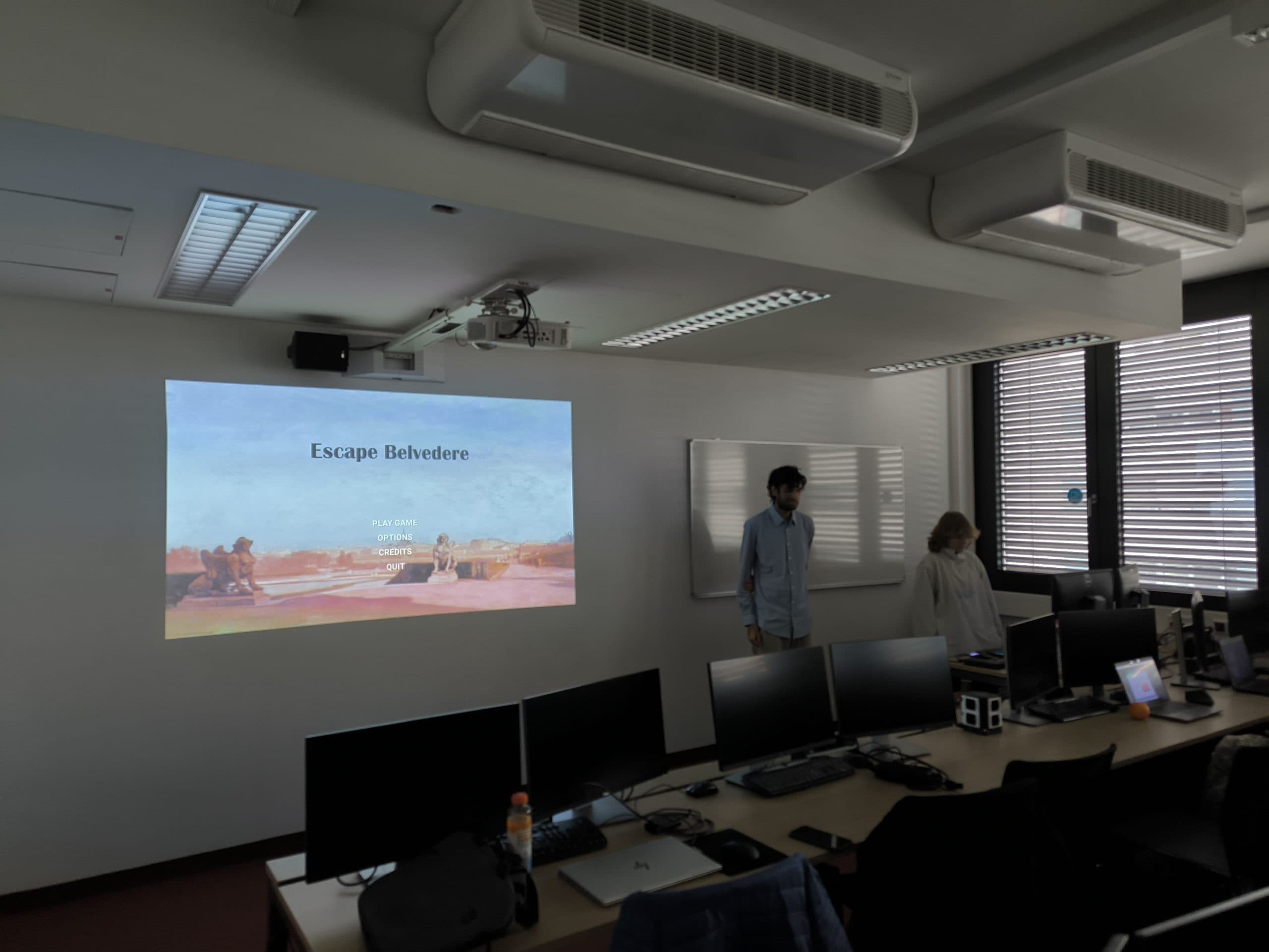 Zwei Personen stehen vorne in einem Klassenzimmer mit Computern auf den Tischen. Auf einem Bildschirm wird 'Escape Belvedere' mit einem gemalten Hintergrund und Spielmenüoptionen angezeigt. Durch die teilweise geschlossenen Jalousien fällt Sonnenlicht ein.