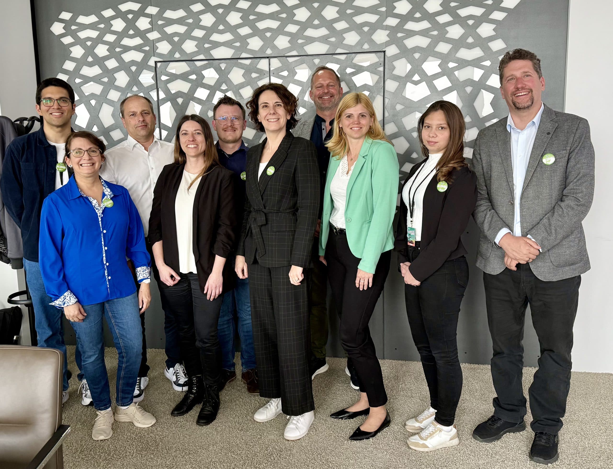 Eine Gruppe von zehn Erwachsenen, Männer und Frauen, steht in einem Innenraum vor einer gemusterten Wand und posiert lächelnd für ein Gruppenfoto. Die meisten von ihnen tragen legere Business-Kleidung und grüne Abzeichen.