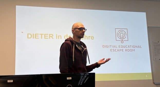 Eine Person steht vor einer projizierten Folie mit den Titeln "DIETER in der Lehre" und "Digital Educational Escape Room" und hält eine Präsentation in einem modernen Klassenzimmer mit Computern und einem Whiteboard.