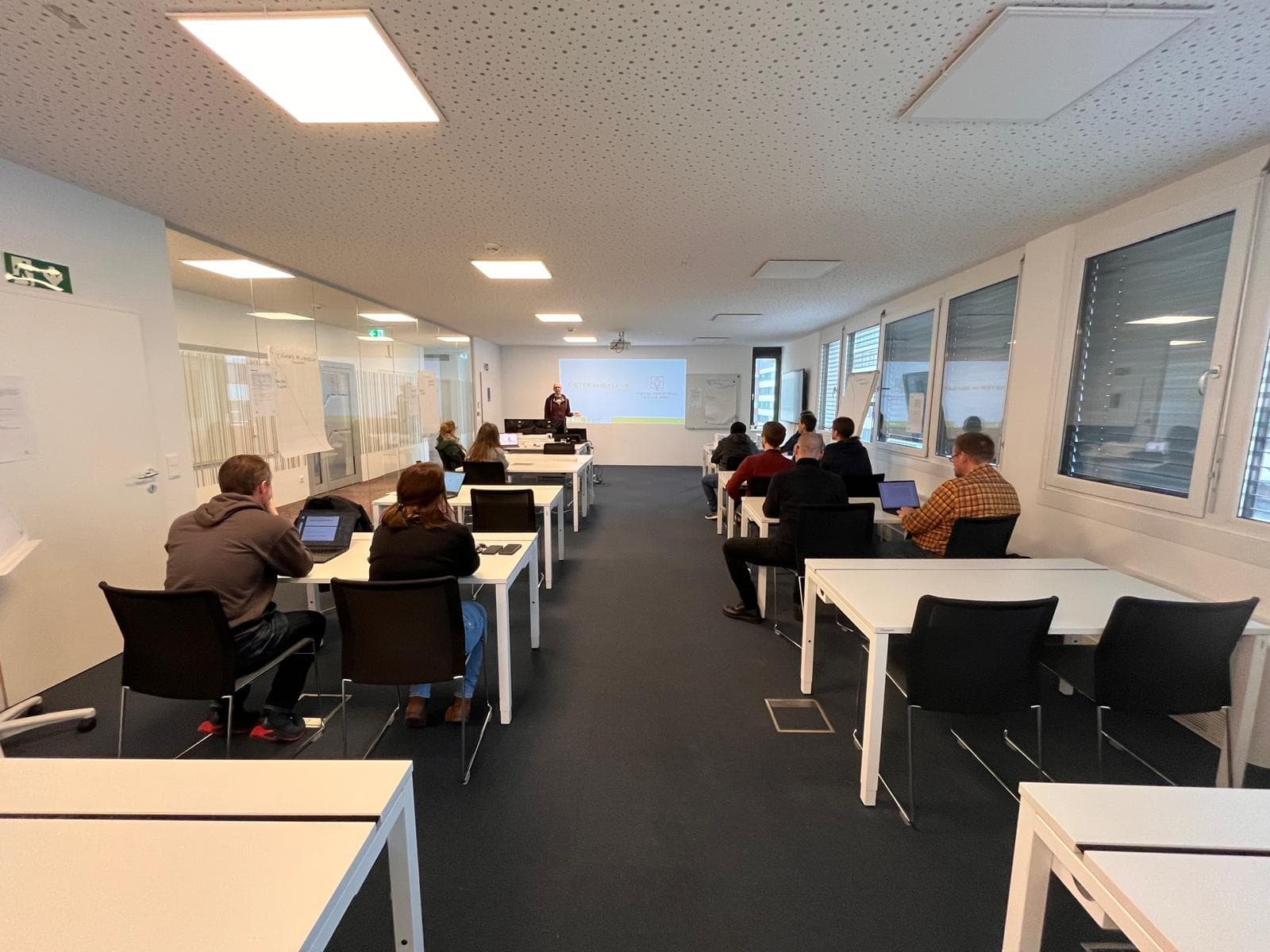 Ein Klassenzimmer mit mehreren Personen, die an Tischen sitzen und einem Vortragenden gegenüberstehen, der eine Präsentation mit Dias auf einer Projektionsfläche hält. Der Raum ist modern und gut beleuchtet mit großen Fenstern.