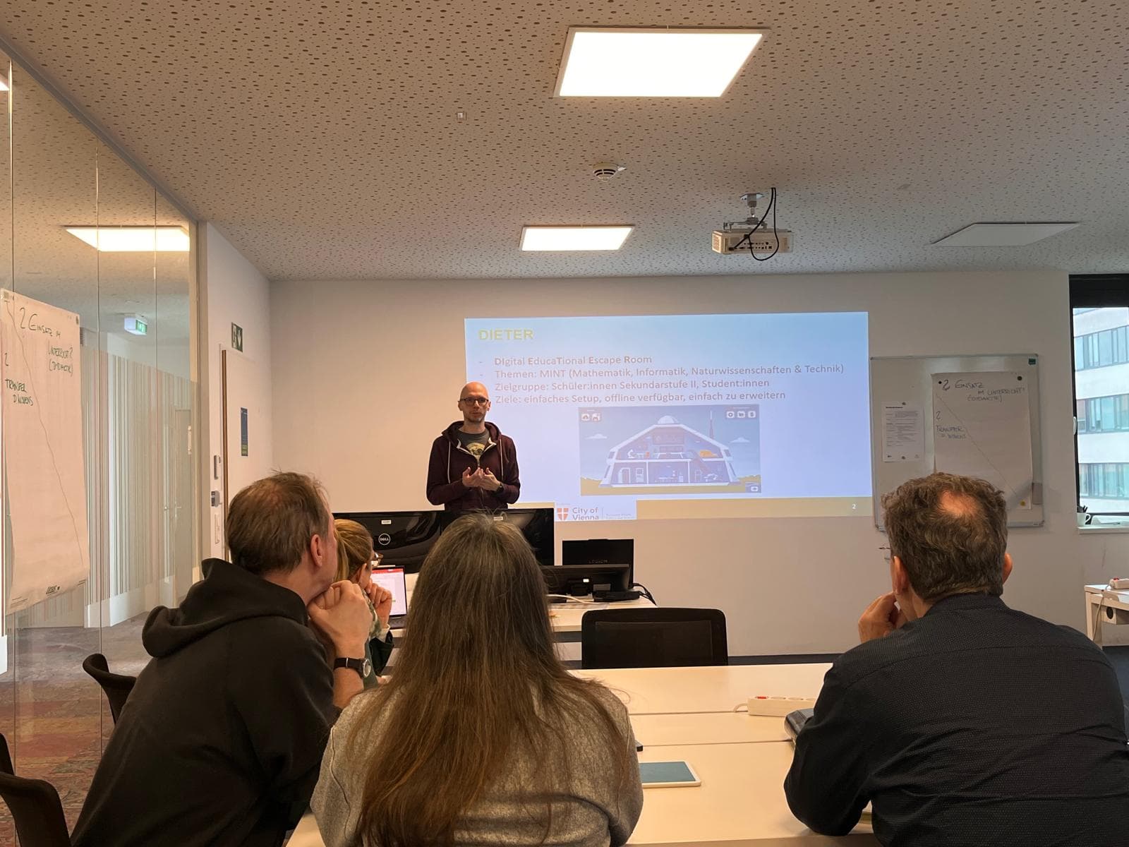 Ein Mann steht vorne in einem Besprechungsraum und hält eine Präsentation vor vier sitzenden Personen. Eine Folie über einen 'Digital Escape Room' wird an die Wand hinter ihm projiziert. Im Raum sind Whiteboards und Büromaterialien zu sehen.