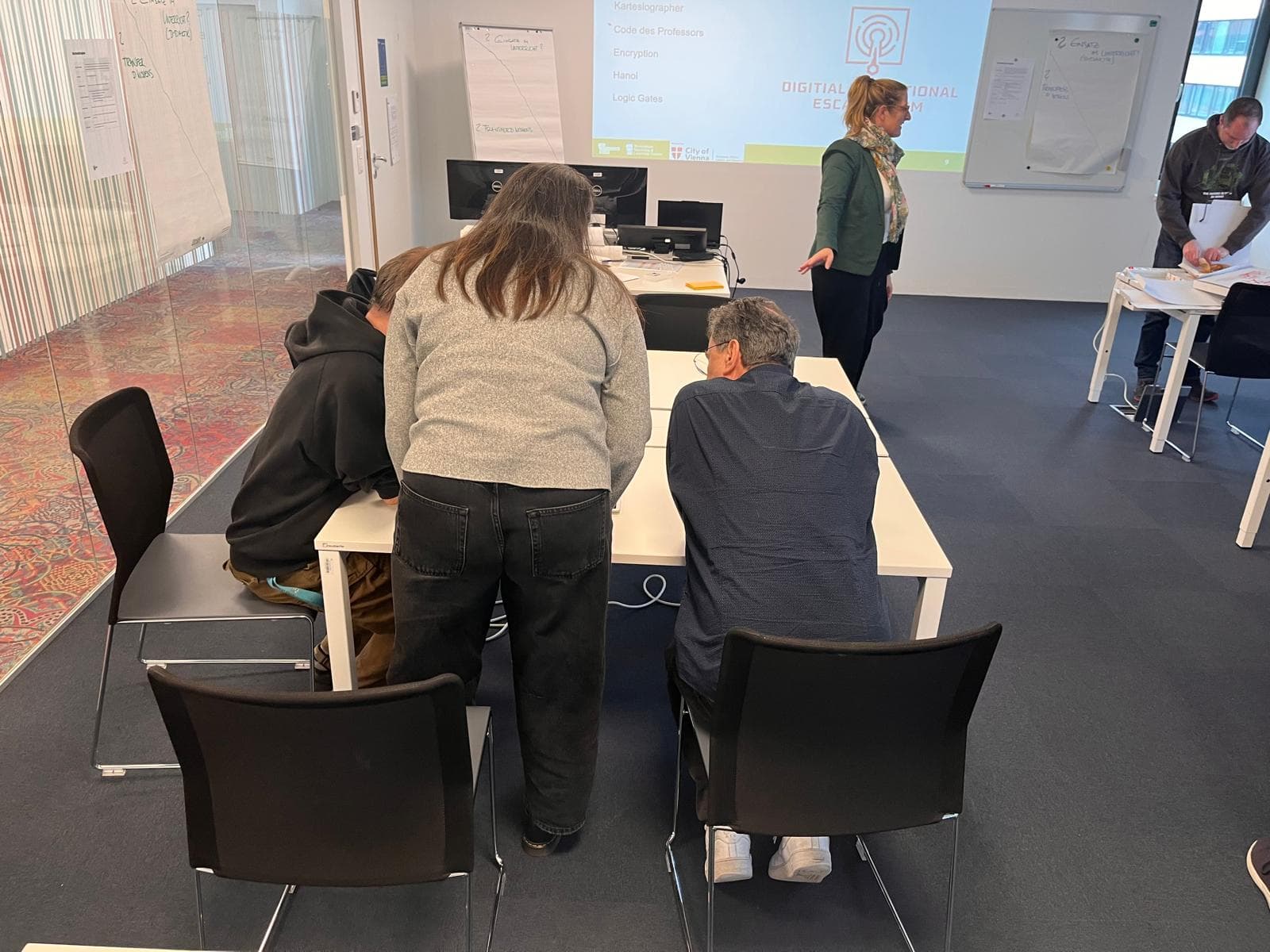 Vier Personen befinden sich in einem modernen Klassenzimmer. Drei sind um einen Tisch versammelt und arbeiten zusammen, während eine Person in der Nähe einer Tafel im vorderen Teil des Raumes steht. Eine weitere Person schreibt an einem anderen Tisch.