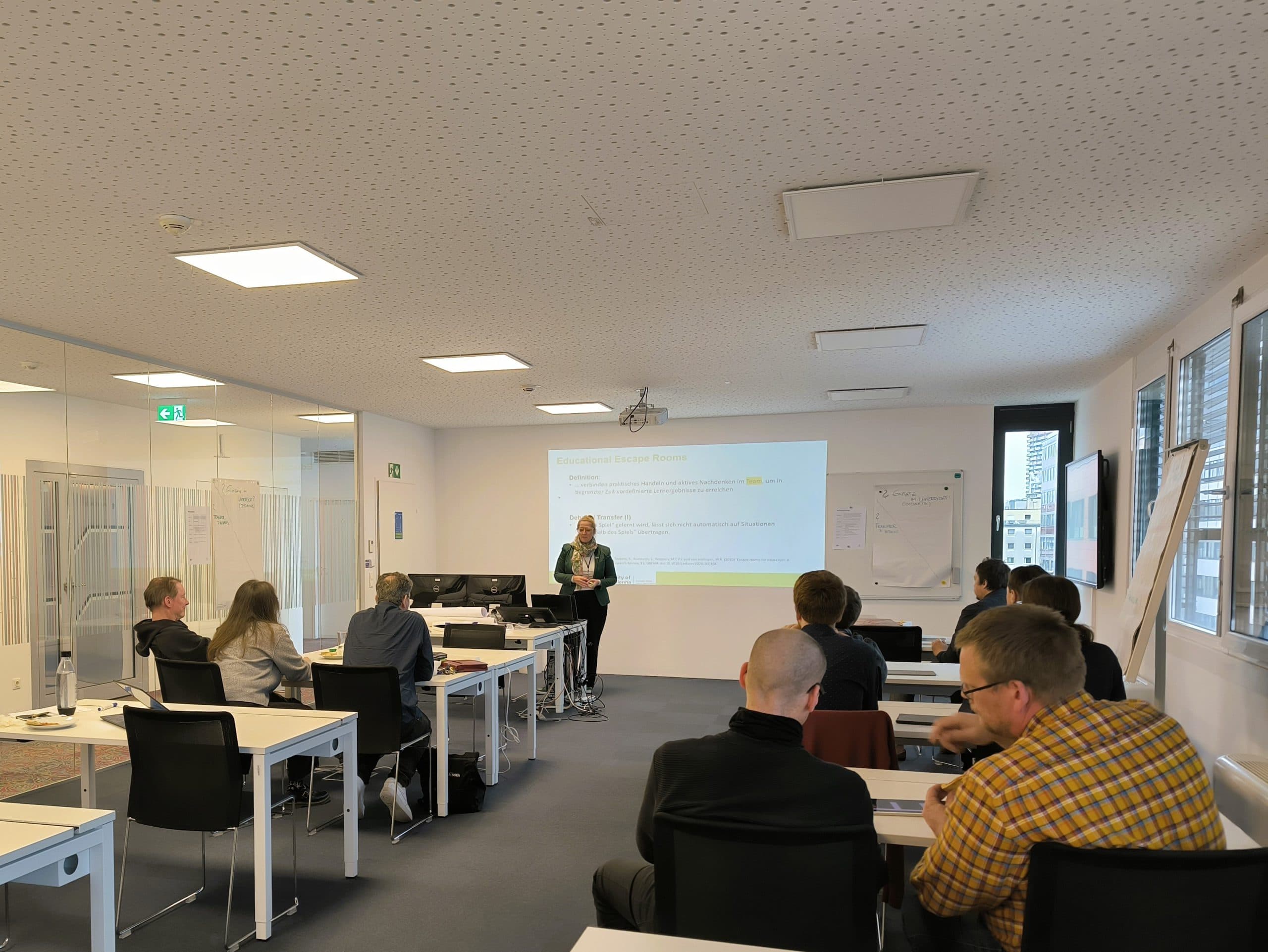 Eine Frau steht vorne in einem modernen Klassenzimmer und hält einer Gruppe von sitzenden Erwachsenen einen Vortrag. Ein großer Bildschirm zeigt Informationen an, und der Raum ist durch mehrere Fenster hell erleuchtet.