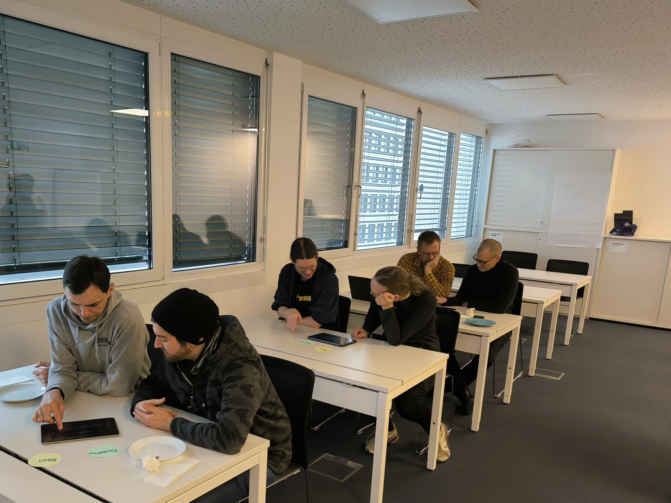 Sechs Personen sitzen paarweise an weißen Tischen in einem hellen Klassenzimmer, jedes Paar schaut auf ein Tablet. Auf den Tischen stehen Teller mit Essen, und große Fenster säumen eine Wand. Die Atmosphäre wirkt konzentriert und kooperativ.