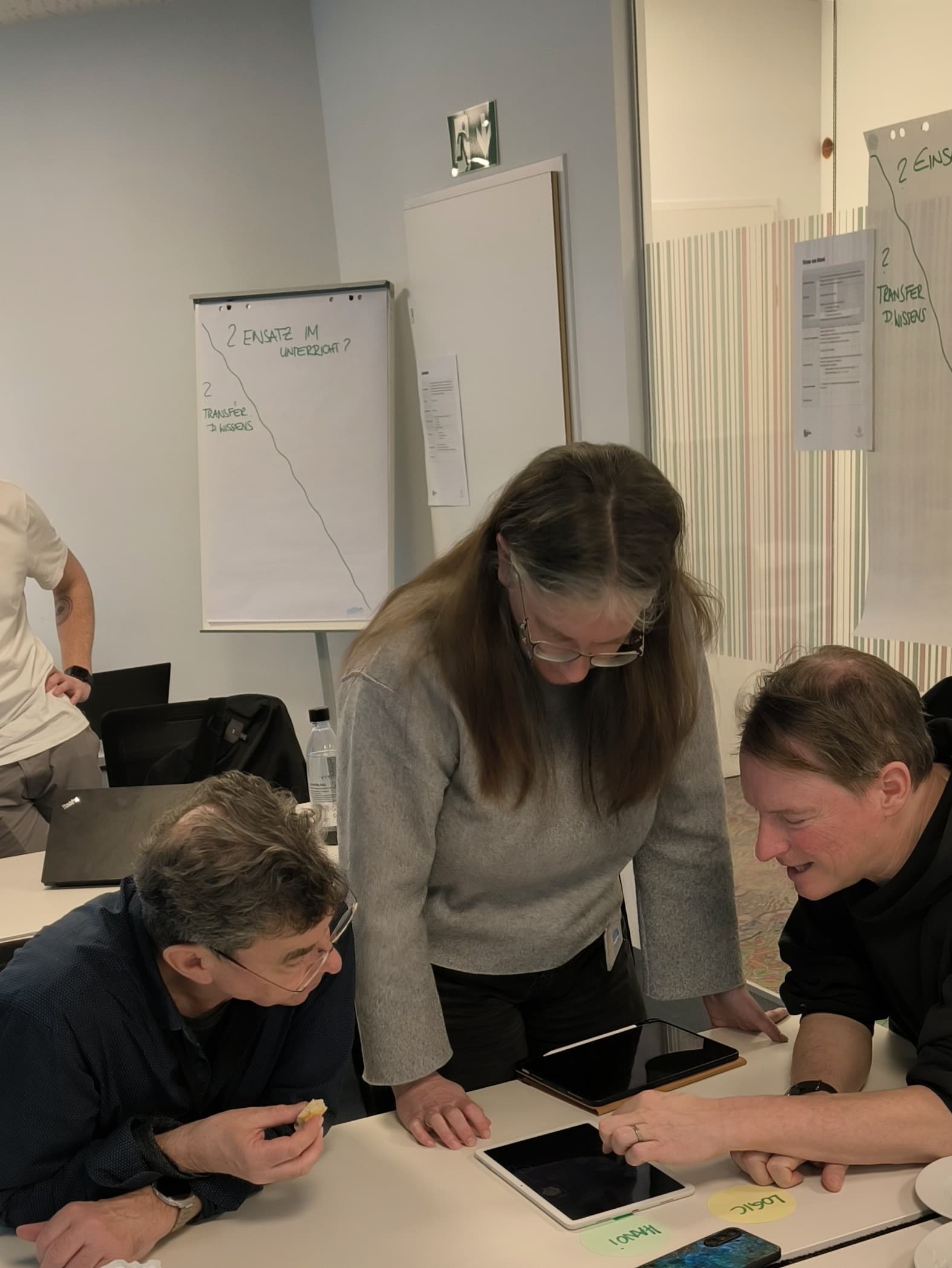 Drei Personen sind um einen Tisch versammelt und schauen gemeinsam auf einen Tablet-Bildschirm. Im Hintergrund sind zwei Flipcharts mit handschriftlichen Notizen zu sehen, die auf eine Zusammenarbeit oder einen Workshop hindeuten.