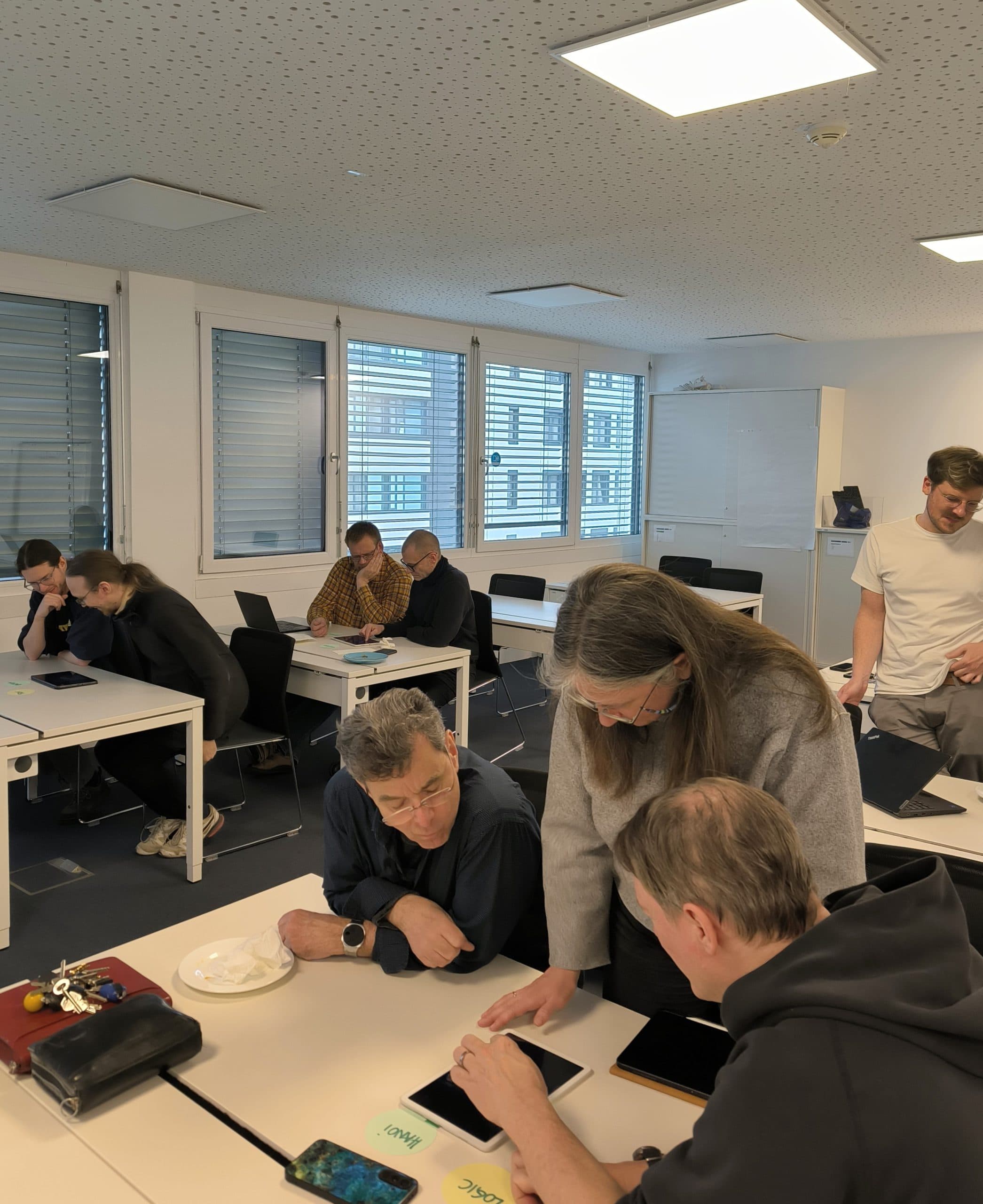 Die Menschen sitzen in Gruppen an Tischen in einem hellen Klassenzimmer und benutzen Tablets. Eine Person steht und hilft anderen. Große Fenster lassen natürliches Licht herein, und mehrere Personen sind in Diskussionen oder Arbeiten vertieft.