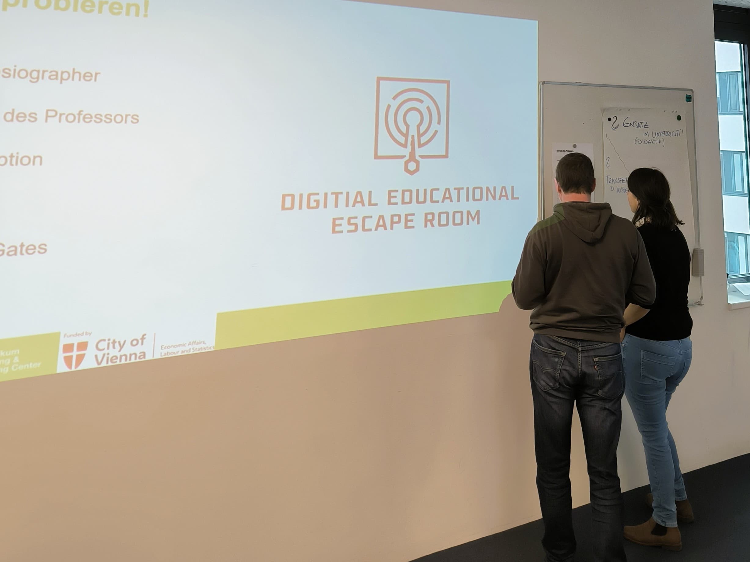 Zwei Personen stehen vor einem Whiteboard, neben einer projizierten Präsentationsfolie mit der Aufschrift Digital Educational Escape Room und einem Logo. Der Raum scheint eine Bildungs- oder Workshopumgebung zu sein.