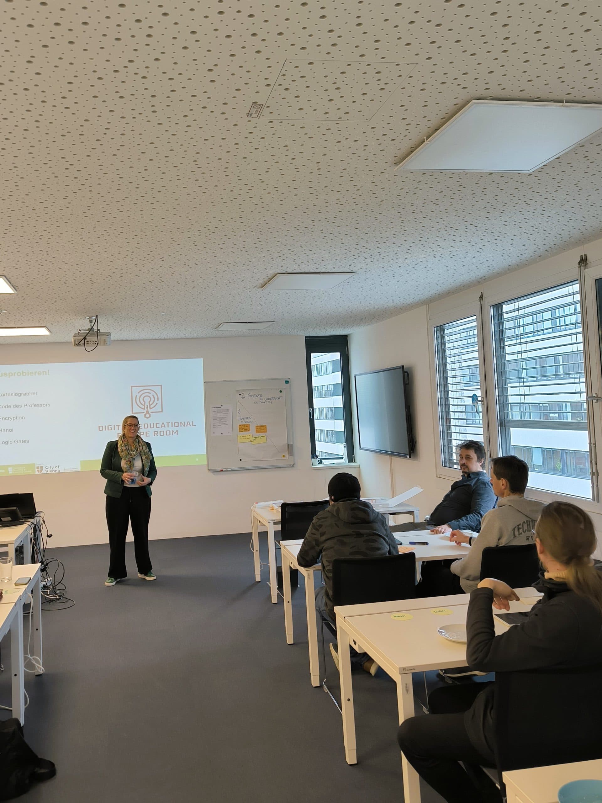 Eine Frau steht vorne in einem Klassenzimmer und hält eine Präsentation vor vier sitzenden Personen. Hinter ihr zeigt ein Projektor ein Dia mit dem Titel Digitaler Bildungsauftrag. Der Raum hat große Fenster und moderne Möbel.