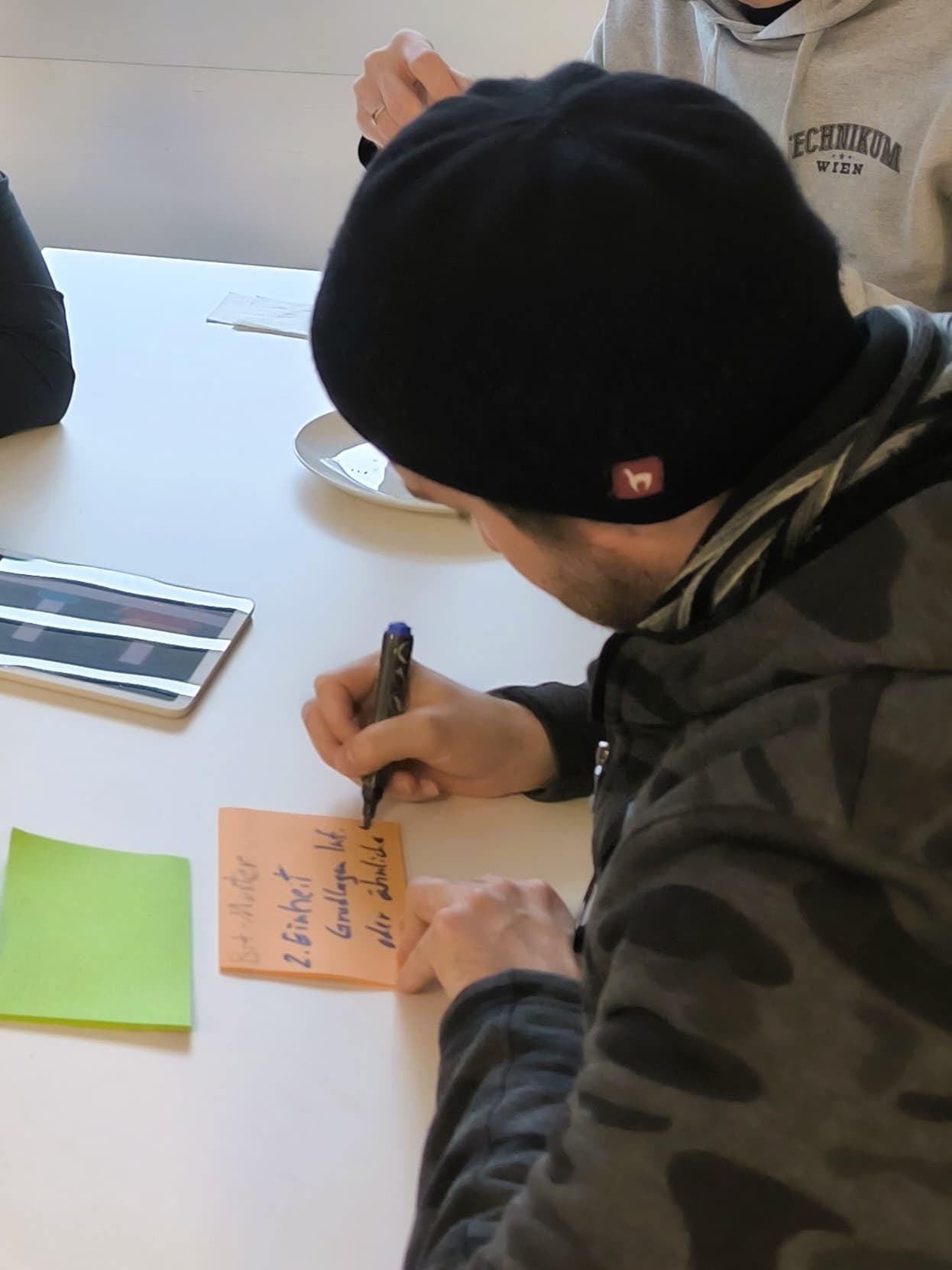 Eine Person, die einen dunklen Kapuzenpulli und eine Mütze trägt, schreibt mit einem schwarzen Stift auf einem braunen Klebezettel an einem weißen Tisch Notizen. Eine grüne Haftnotiz, ein Tablet und der Arm einer anderen Person sind ebenfalls zu sehen.