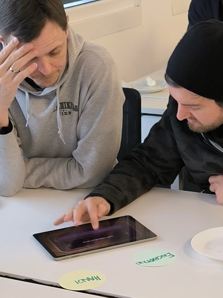 Zwei Personen sitzen an einem Tisch, konzentriert auf ein Tablet-Gerät. Einer berührt den Bildschirm, während der andere nachdenklich zusieht. Auf dem Tisch daneben liegen runde Papierschilder mit der Aufschrift ENERGIZE! und HELLO!.