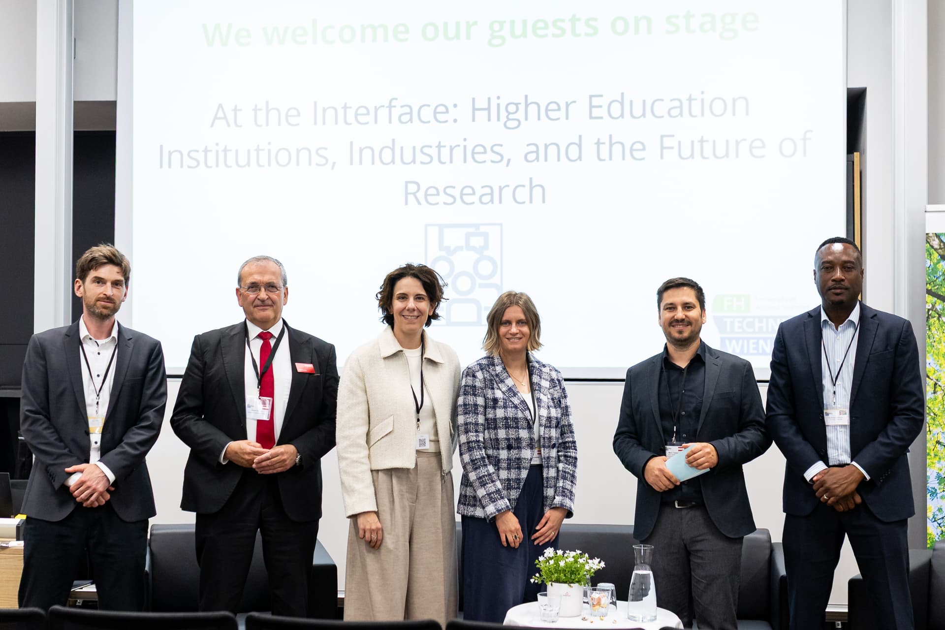 Sechs professionell gekleidete Personen stehen vor einem Bildschirm, auf dem der Text angezeigt wird: An der Schnittstelle: Hochschuleinrichtungen, Industrie und die Zukunft der Forschung.