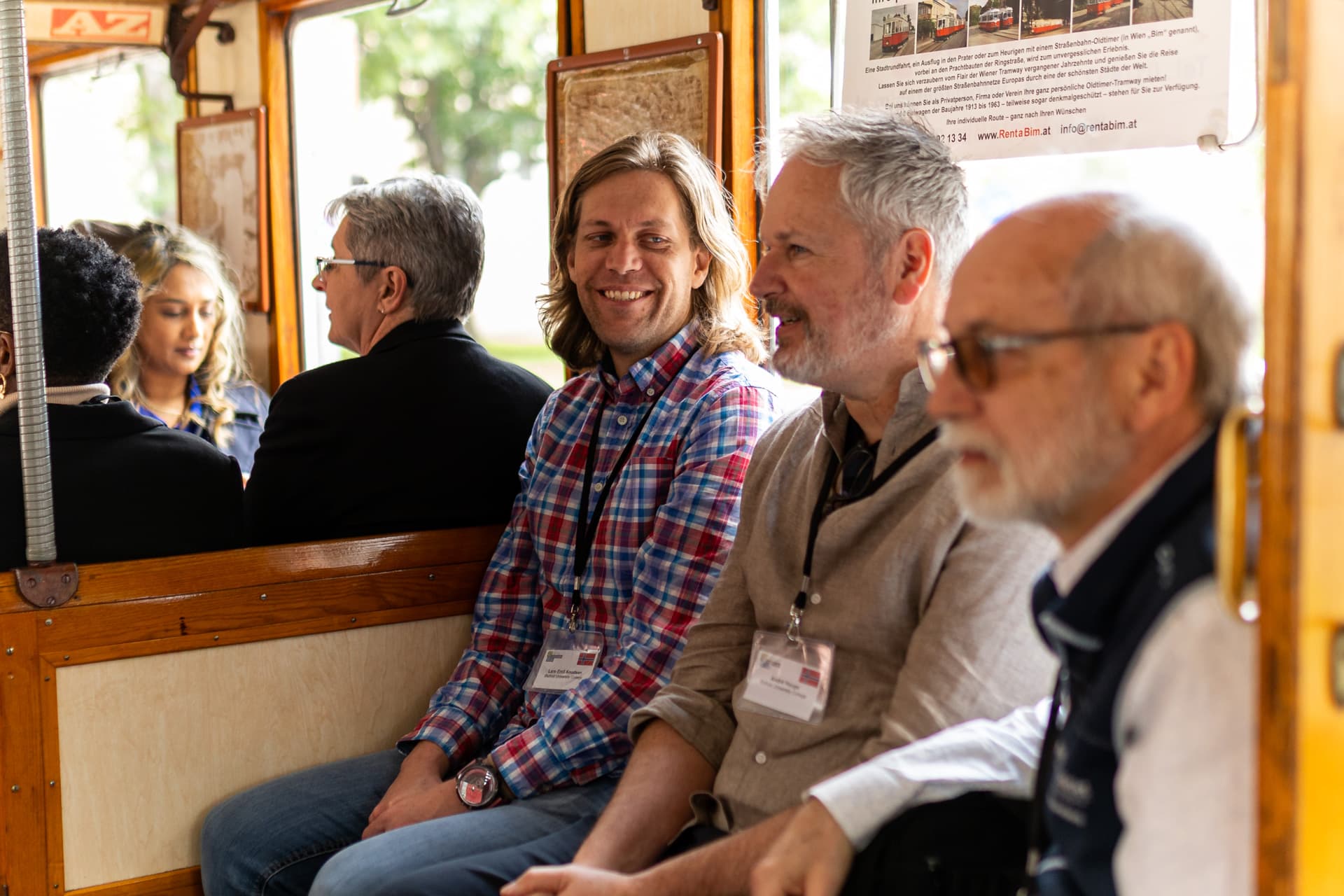 Vier Personen sitzen und unterhalten sich in einem hölzernen Wagen oder einer Straßenbahn. Zwei Männer im Vordergrund sind lächelnd im Fokus, während andere Fahrgäste im Hintergrund zu sehen sind. Das Sonnenlicht scheint durch die Fenster.