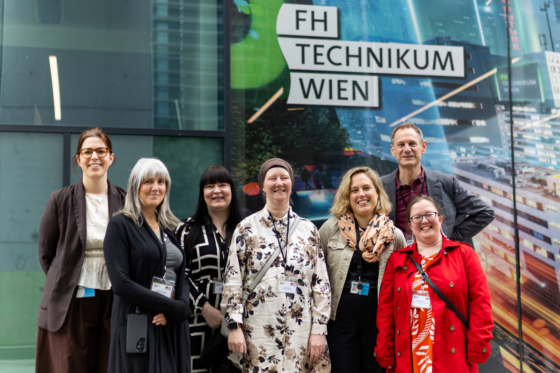 Eine Gruppe von sieben lächelnden Erwachsenen posiert vor einem Glasgebäude mit einem Schild, auf dem FH Technikum Wien steht. Sie tragen Konferenzabzeichen und verschiedene Kleidungsstile.