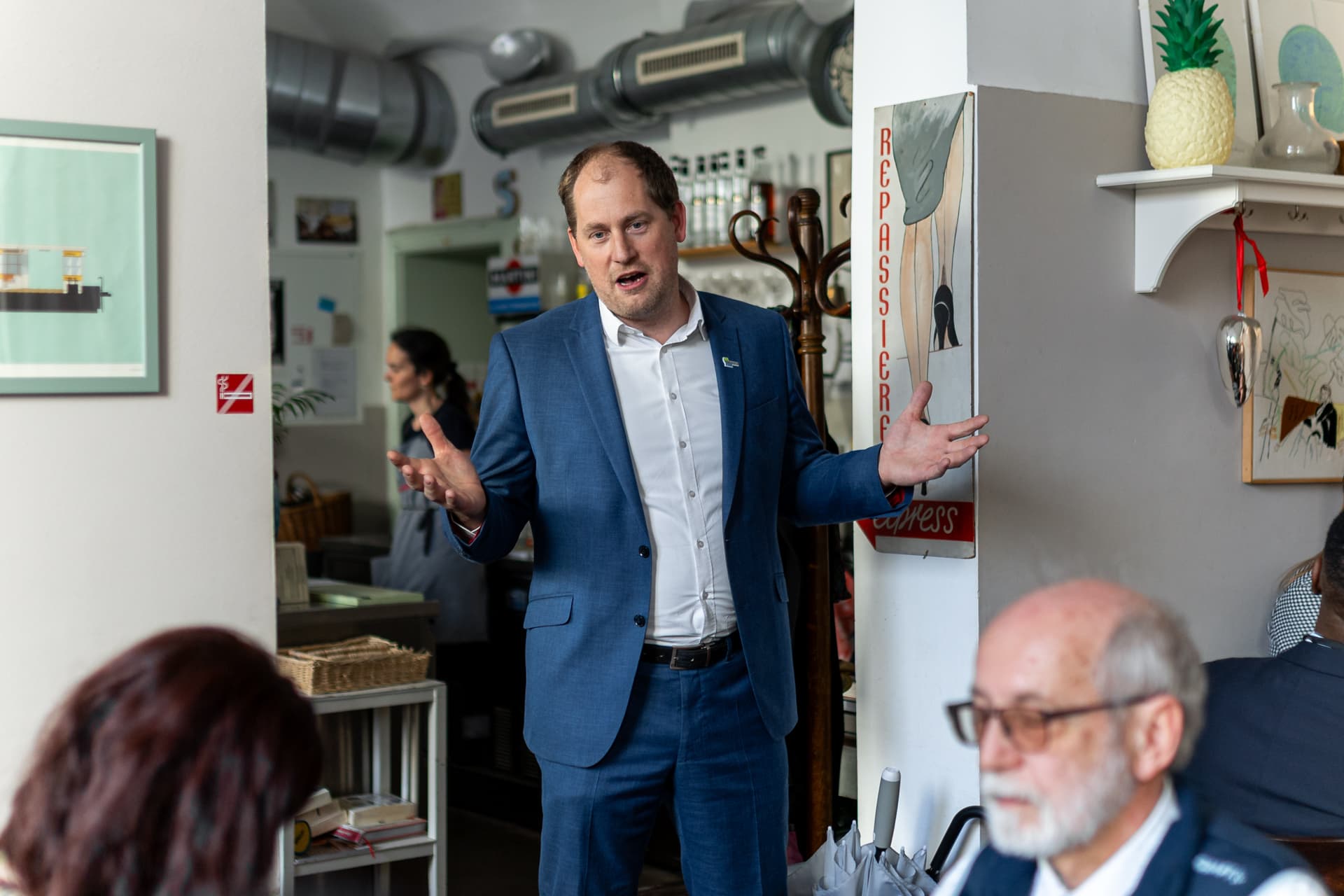 Ein Mann in einem blauen Anzug steht in einem Raum und spricht mit offenen Händen zu einer kleinen Gruppe. Es handelt sich um einen legeren Raum mit Kunst an den Wänden, in dem mehrere Personen sitzen und zuhören.