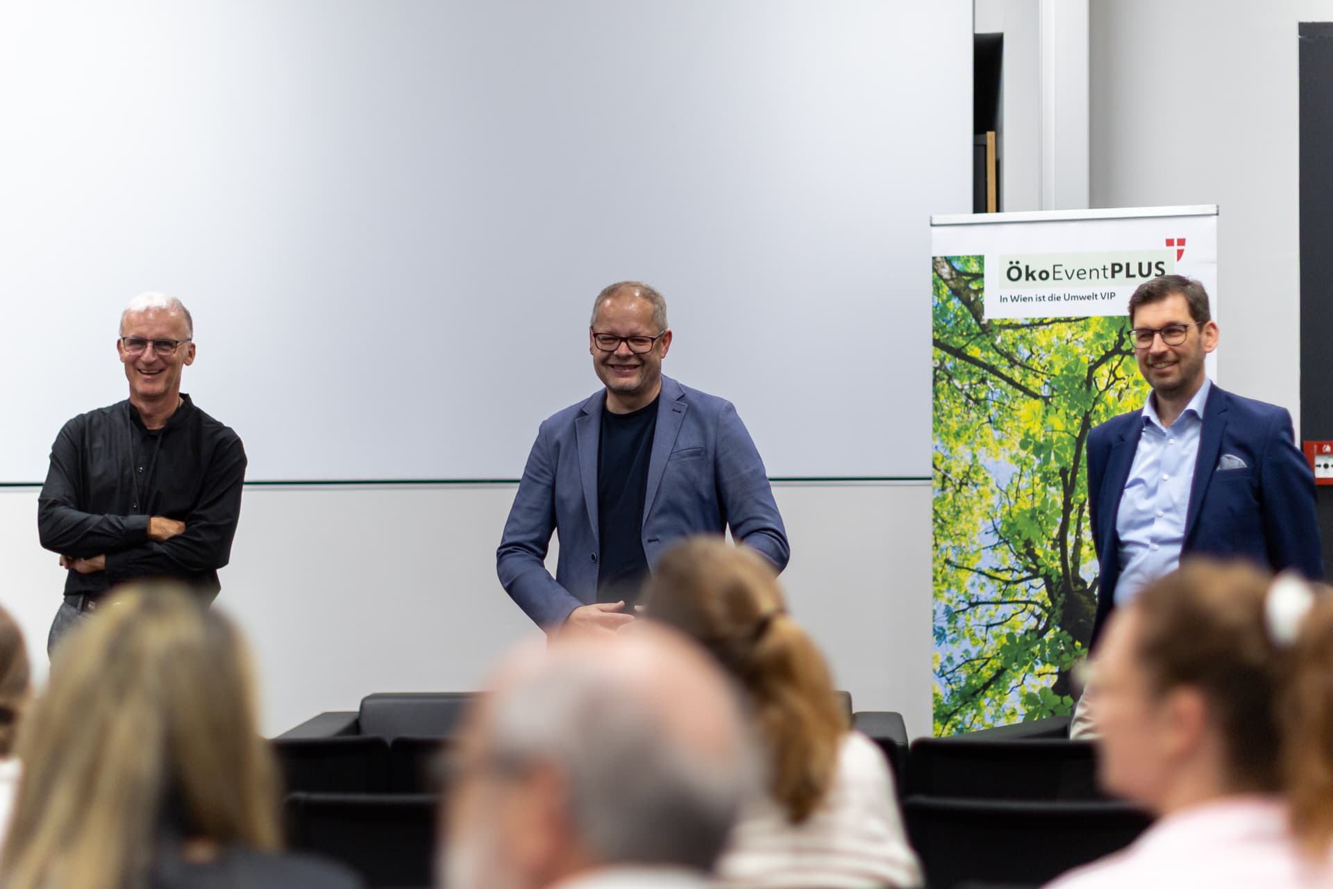 Drei Männer stehen vorne in einem Raum und blicken lächelnd in die Runde. Hinter ihnen ist ein Banner mit der Aufschrift ÖkoEventPLUS und einem Bild mit grünen Ästen zu sehen. Die Umgebung scheint eine Konferenz oder ein Seminar zu sein.