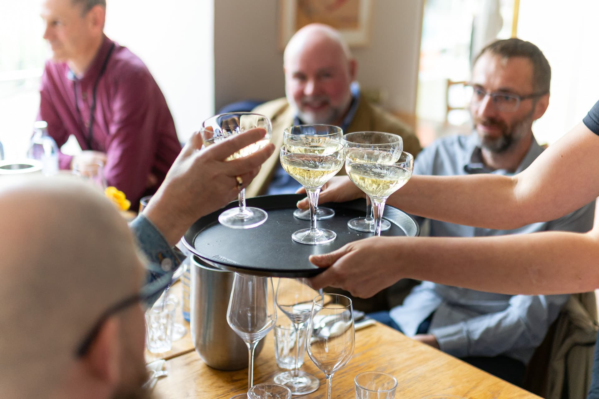 Eine Gruppe von Menschen sitzt um einen Tisch herum und lächelt, während jemand Gläser mit Weißwein auf einem Tablett serviert. Leere Weingläser und ein Weinkühler stehen ebenfalls auf dem Holztisch.