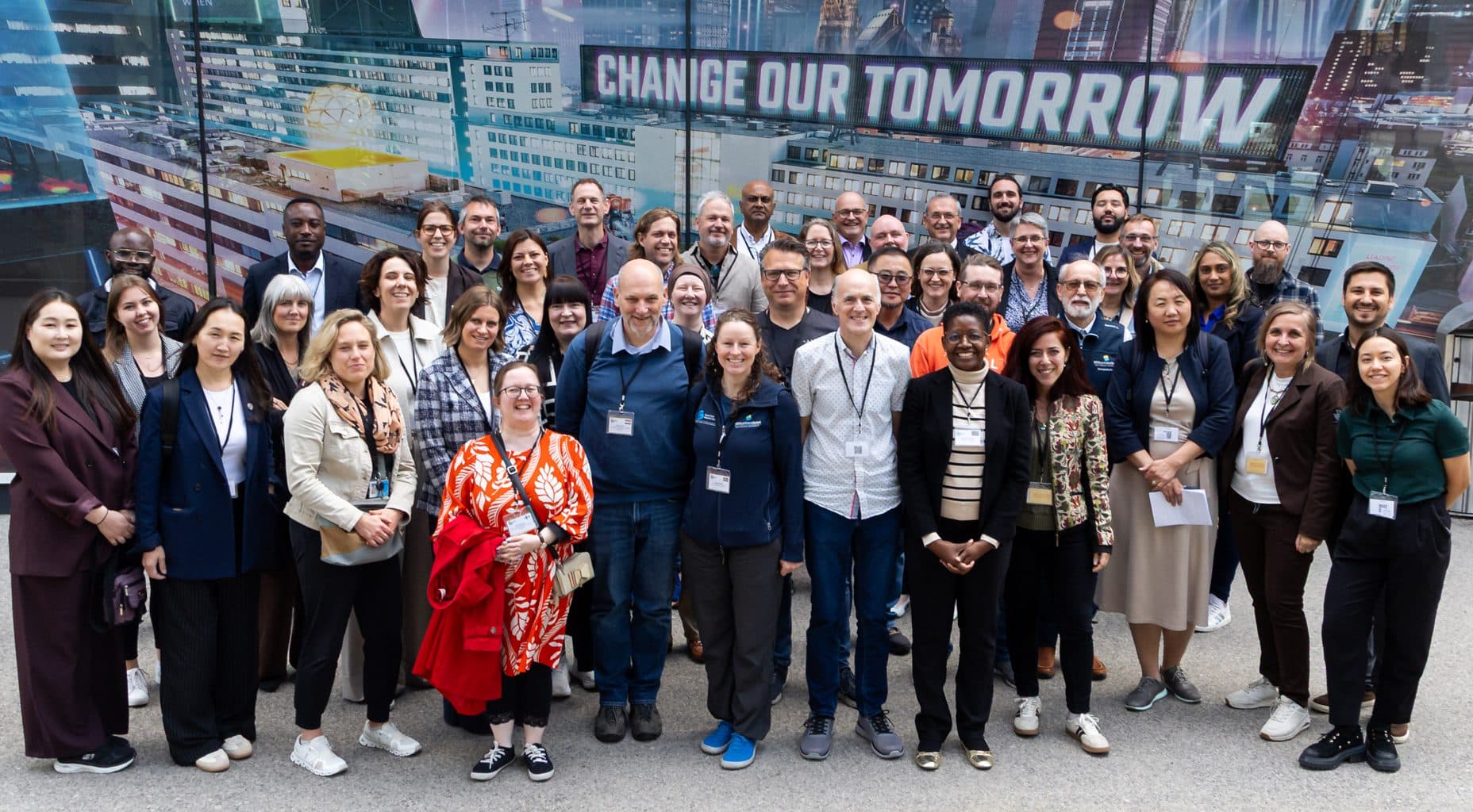 Eine bunt gemischte Gruppe von Menschen steht im Freien zusammen und lächelt in die Kamera vor einem farbenfrohen städtischen Wandgemälde mit der Aufschrift "Change Our Tomorrow" im Hintergrund.