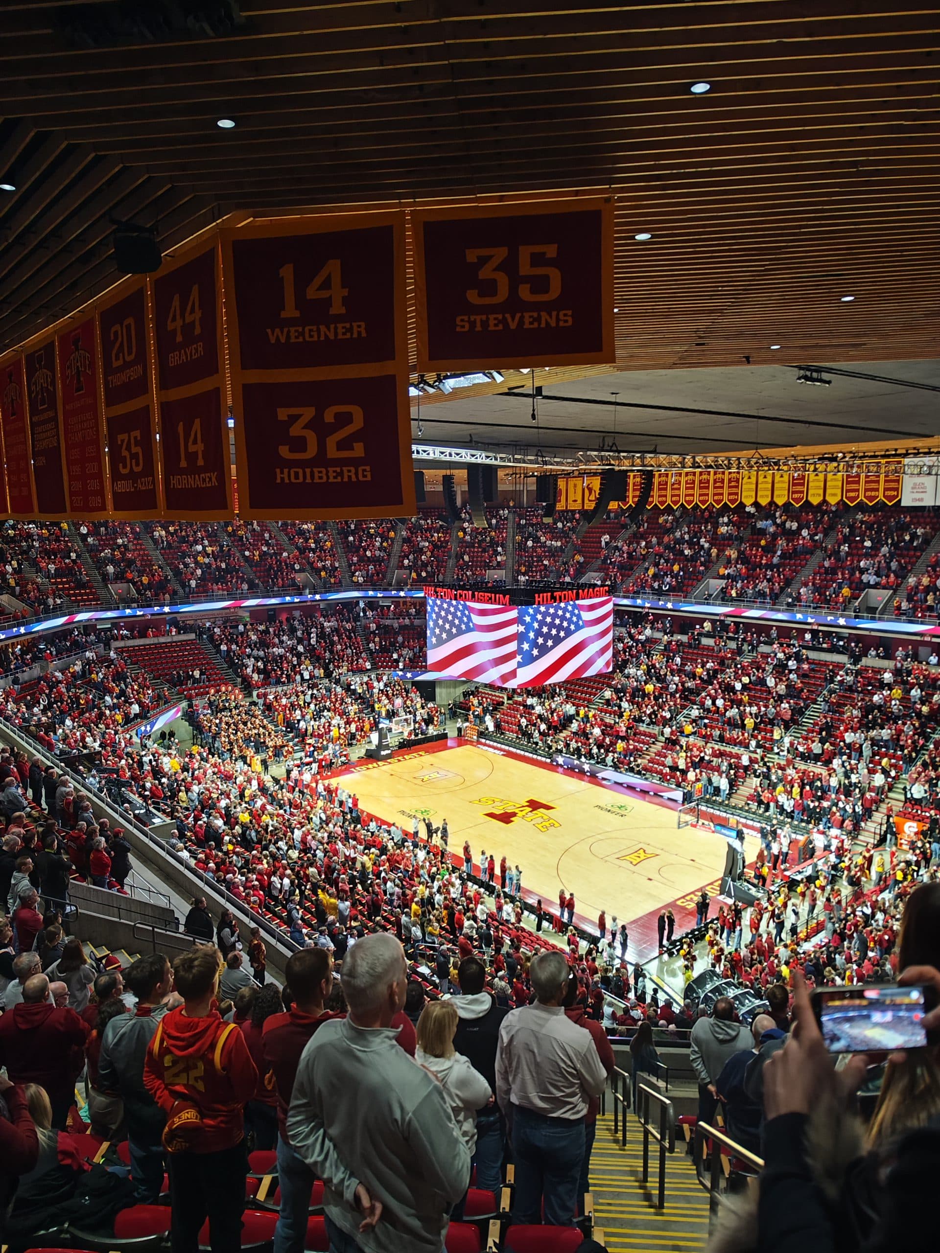Eine große Menschenmenge steht in einer Basketball-Arena, während eine riesige amerikanische Flagge auf dem Spielfeld und der Anzeigetafel zu sehen ist, wobei die meisten Menschen der Flagge zugewandt sind. Banner und rote Sitze füllen das Stadion. Einige Leute machen Fotos.