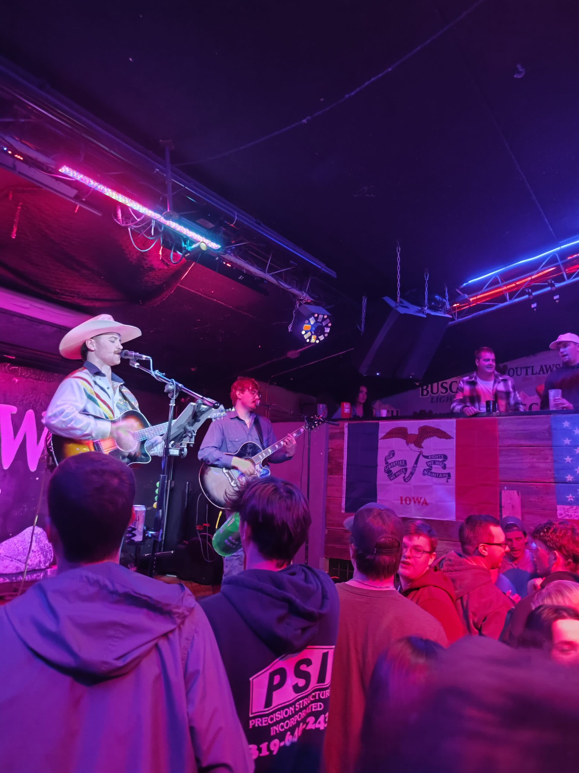 Eine Band tritt auf einer kleinen, neonbeleuchteten Bühne vor einer Menschenmenge auf. Der Leadsänger, der einen Cowboyhut trägt, spielt Gitarre und singt in ein Mikrofon. Das Publikum schaut vom Boden und von einem mit Plakaten geschmückten Balkon aus zu.