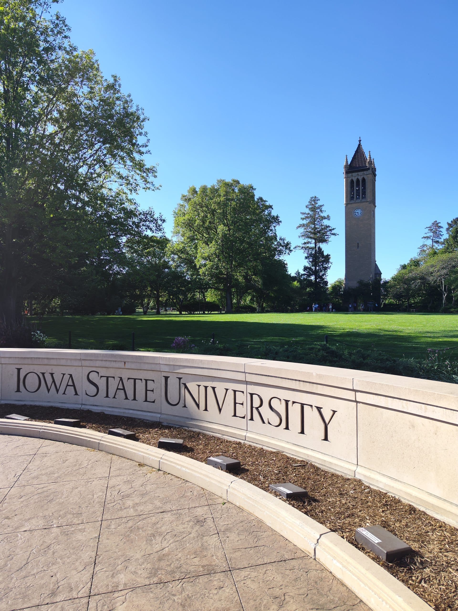 Ein steinernes Schild mit der Aufschrift Iowa State University steht vor einer großen, grasbewachsenen Fläche mit hohen Bäumen und dem Glockenturm des Campanile im Hintergrund unter einem klaren blauen Himmel.