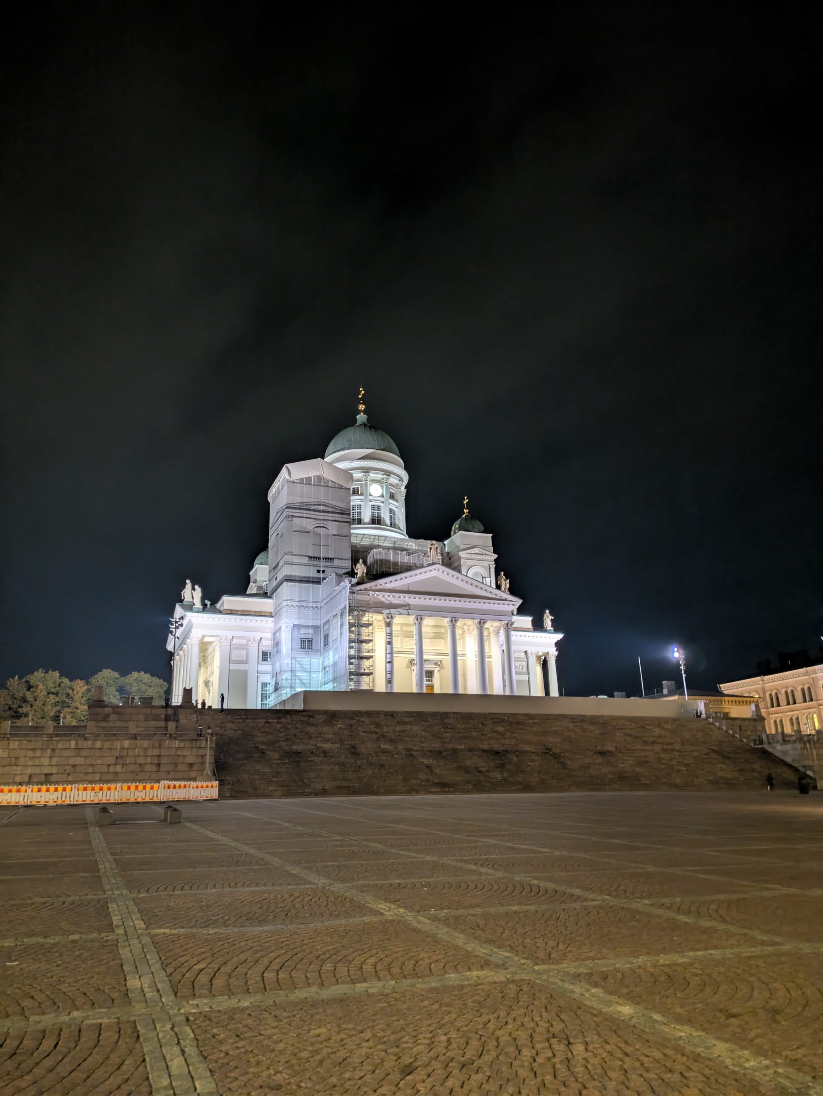 Eine weiße neoklassizistische Kathedrale mit einer großen zentralen Kuppel ist nachts hell erleuchtet und steht auf einer erhöhten Plattform mit breiten Steinstufen vor einem dunklen Himmel und einem leeren Kopfsteinpflasterplatz im Vordergrund.