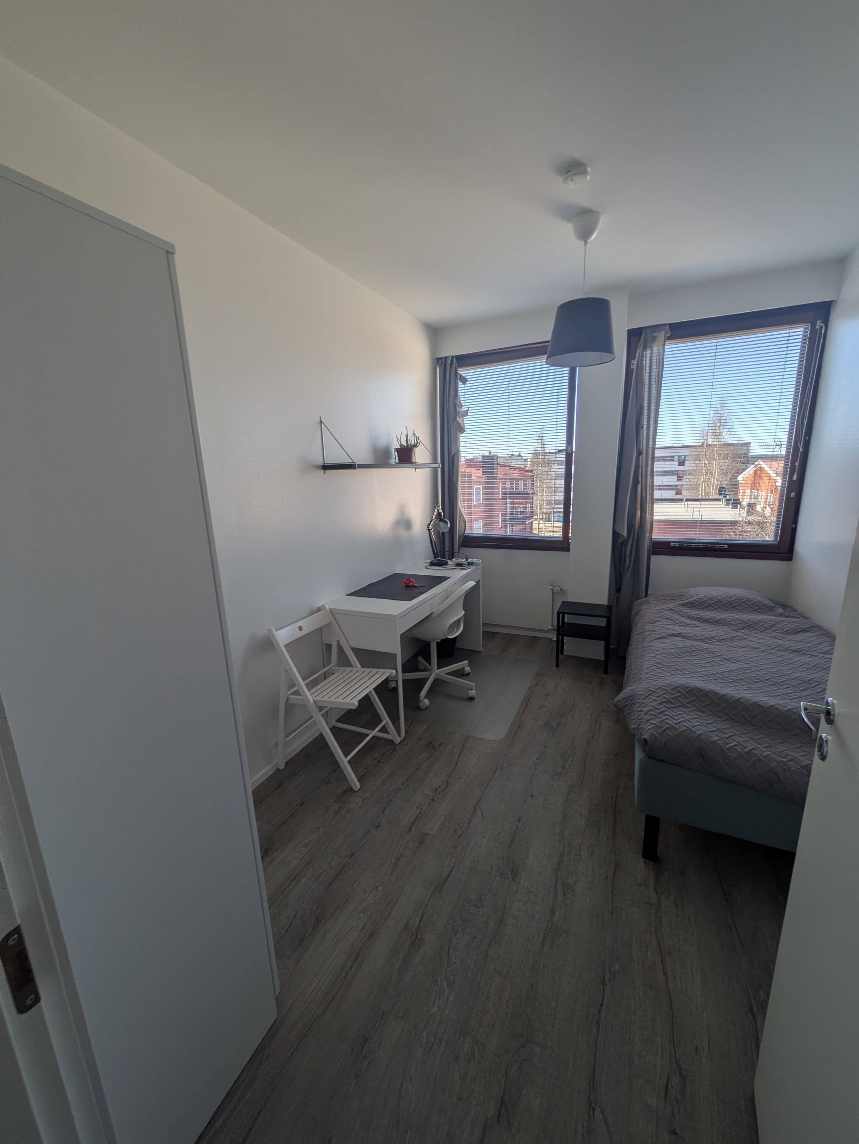 Ein kleines, aufgeräumtes Zimmer mit einem Bett, einem weißen Schreibtisch und Stuhl, Regalen, einem Kleiderschrank und einem Fenster, das Tageslicht hereinlässt. Der Boden ist aus Holz und das Zimmer ist minimalistisch eingerichtet.
