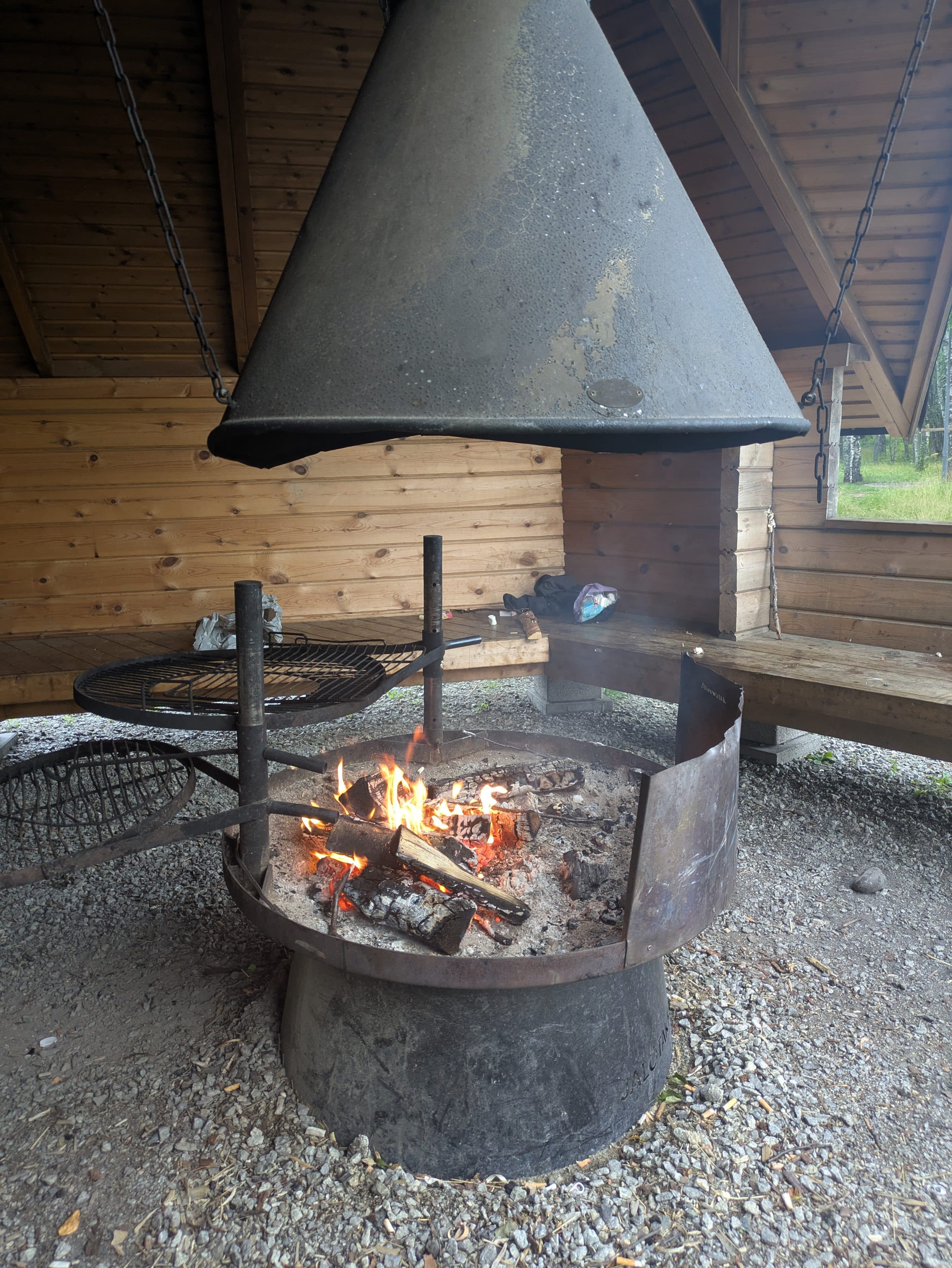 Eine metallene Feuerstelle mit brennendem Holz ist unter einer Holzkonstruktion geschützt. Über dem Feuer hängt eine große Metallhaube, und an den Wänden stehen Bänke. Der Bereich hat eine rustikale Atmosphäre im Freien.