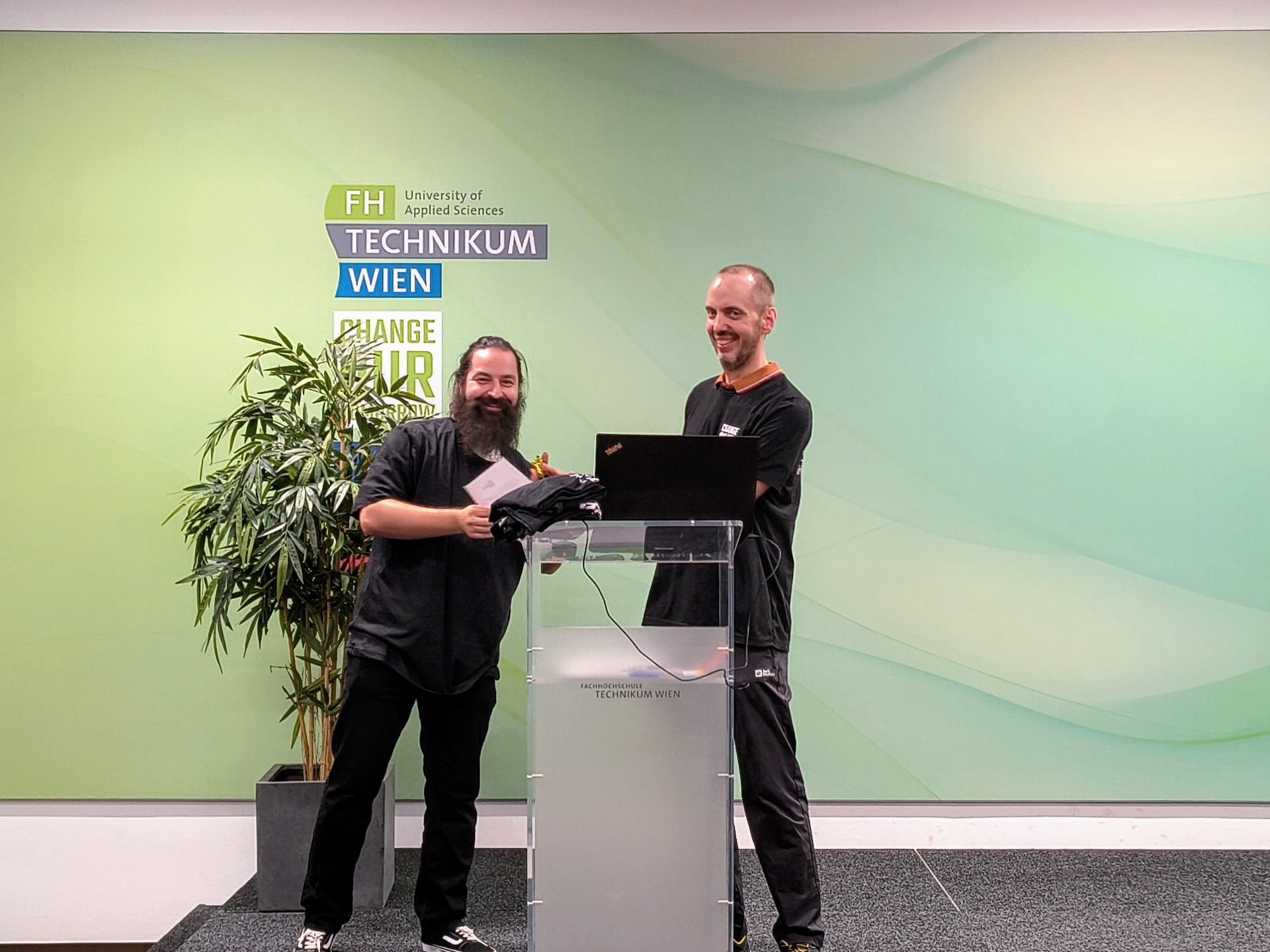 Zwei Männer stehen lächelnd auf einer Bühne neben einem durchsichtigen Rednerpult. Einer hält ein kleines Geschenk in der Hand. Hinter ihnen stehen eine große Pflanze und eine Wand, auf der das Logo „FH Technikum Wien“ sowie der Schriftzug „Change Your Life“ zu sehen sind.