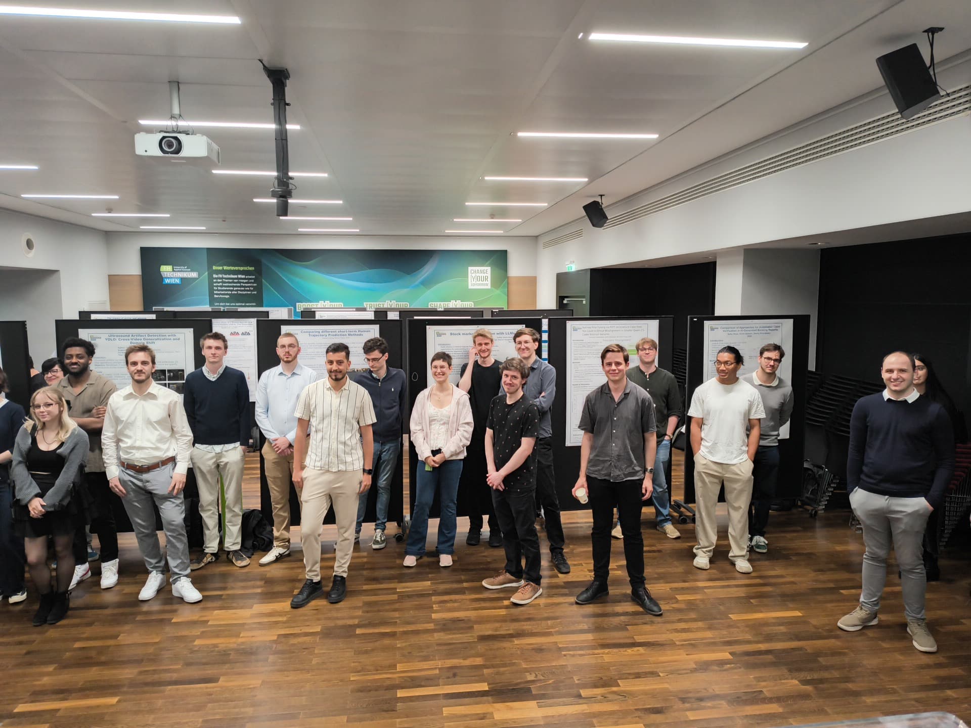 Eine Gruppe von Menschen posiert und lächelt für ein Foto in einem modernen Innenraum mit Holzböden und hinter ihnen aufgehängten Postern. Der Raum ist hell beleuchtet und strahlt eine gepflegte, professionelle Atmosphäre aus.