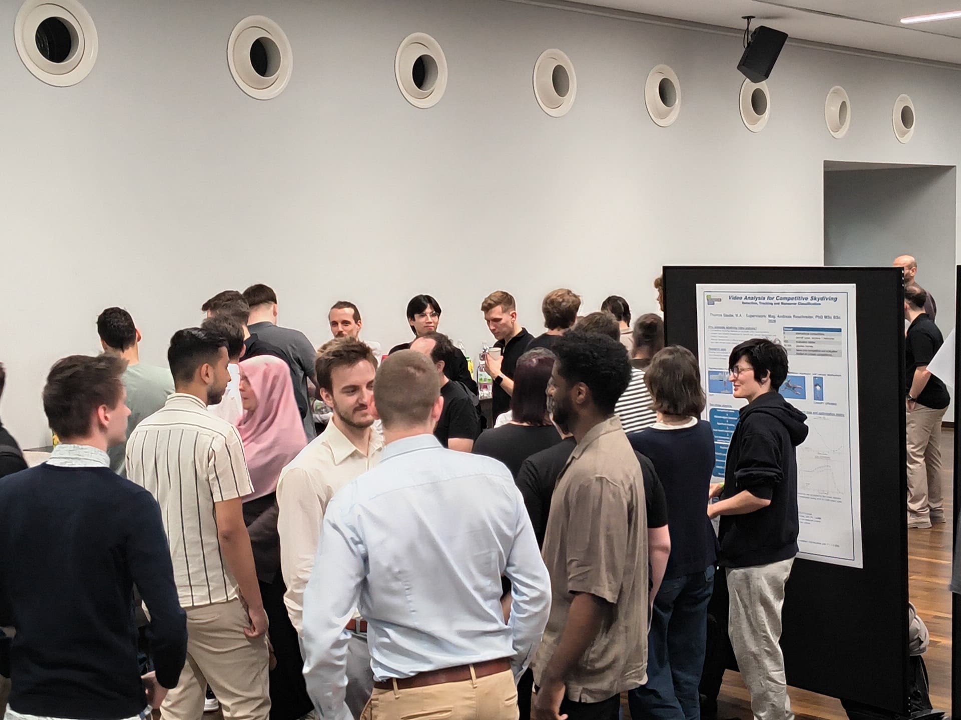 In einem Konferenzraum versammelt sich eine Gruppe von Menschen um eine große Anschlagtafel mit Grafiken und Text und diskutiert. Der Raum verfügt über runde Wandlüftungsöffnungen und Holzböden.