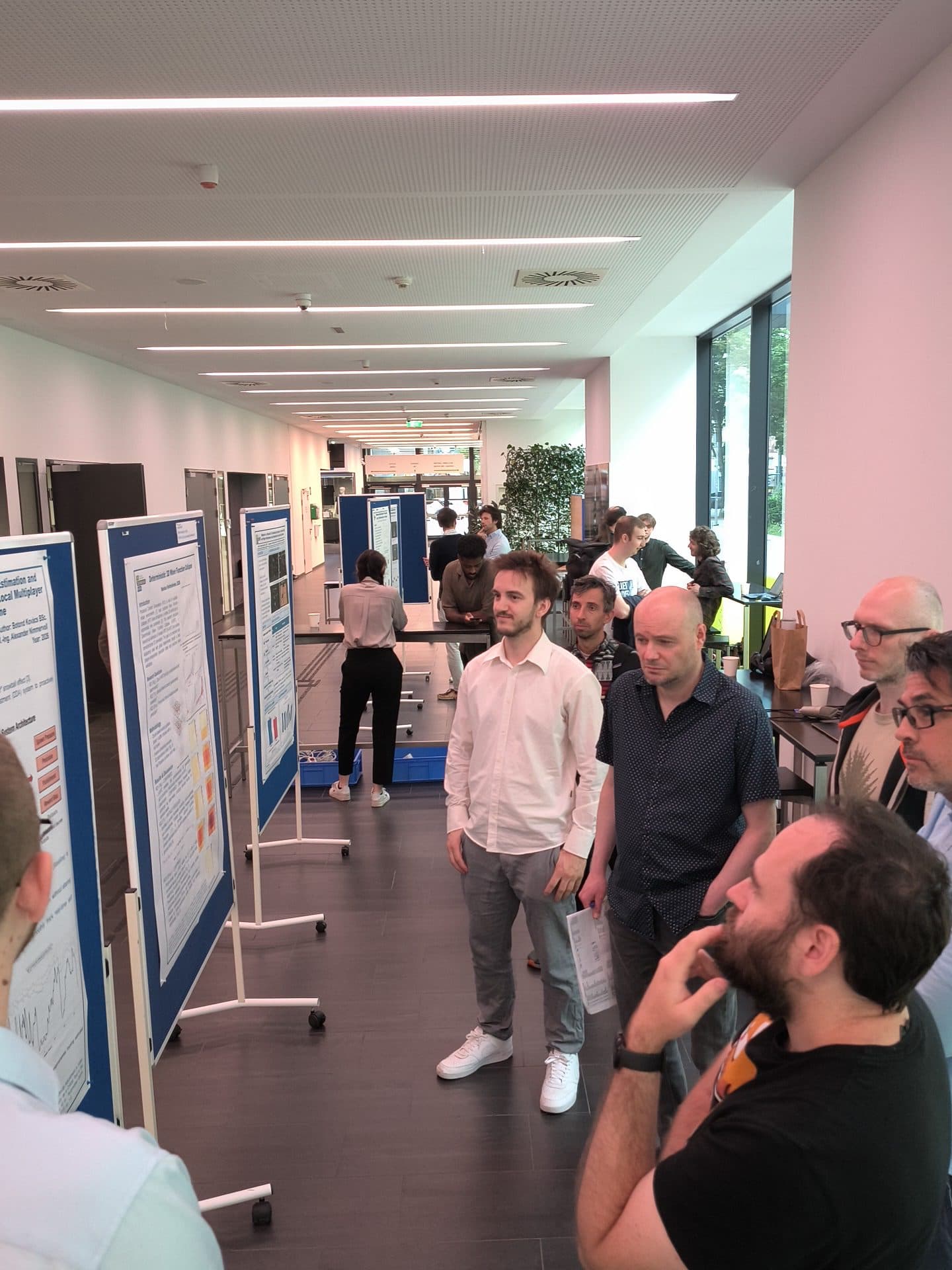 Menschen stehen in einem Flur, betrachten wissenschaftliche Poster, die auf blauen Tafeln ausgestellt sind, und diskutieren darüber. Die Kulisse lässt auf eine akademische oder fachliche Veranstaltung schließen, die von Tageslicht aus großen Fenstern erhellt wird.