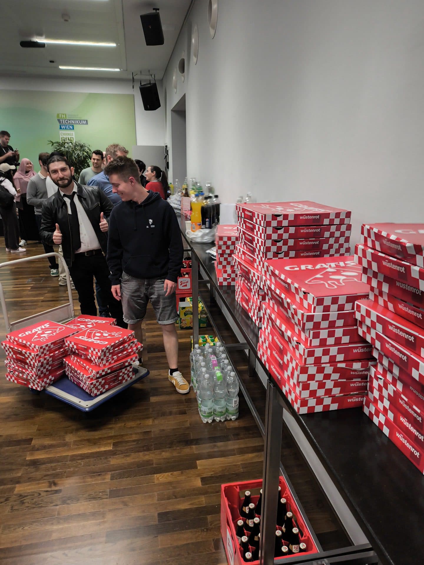 Menschen versammeln sich in einem Raum mit Holzboden. Zwei Männer stehen neben einem Tisch, auf dem rote und weiße Pizzakartons, Getränkeflaschen und Kisten gestapelt sind. Ein Mann streckt den Daumen nach oben, während andere im Hintergrund stehen.