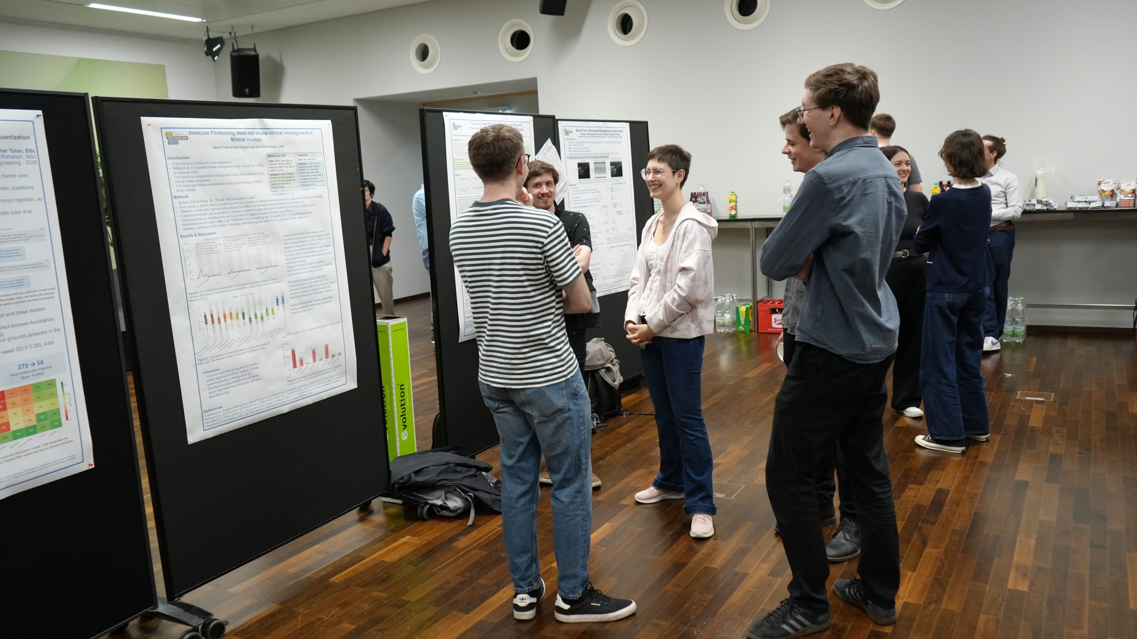 In einem hellen Raum mit Holzboden stehen die Leute in kleinen Gruppen beisammen und diskutieren über wissenschaftliche Poster, die an schwarzen Tafeln angebracht sind. Im Hintergrund sind auf einem Tisch Snacks und Getränke zu sehen.