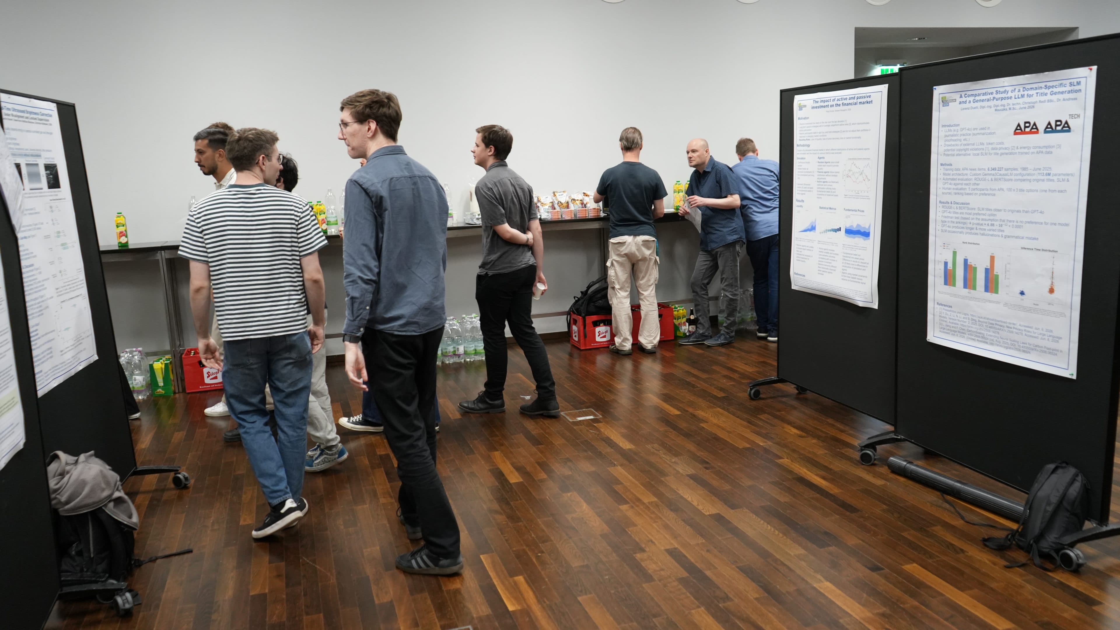 Mehrere Personen stehen und gehen in einem Raum umher, in dem wissenschaftliche Poster an schwarzen Tafeln ausgestellt sind. Einige betrachten die Poster, während sich andere an einem Erfrischungstisch mit Getränken und Snacks versammeln. Der Boden ist aus Holz.