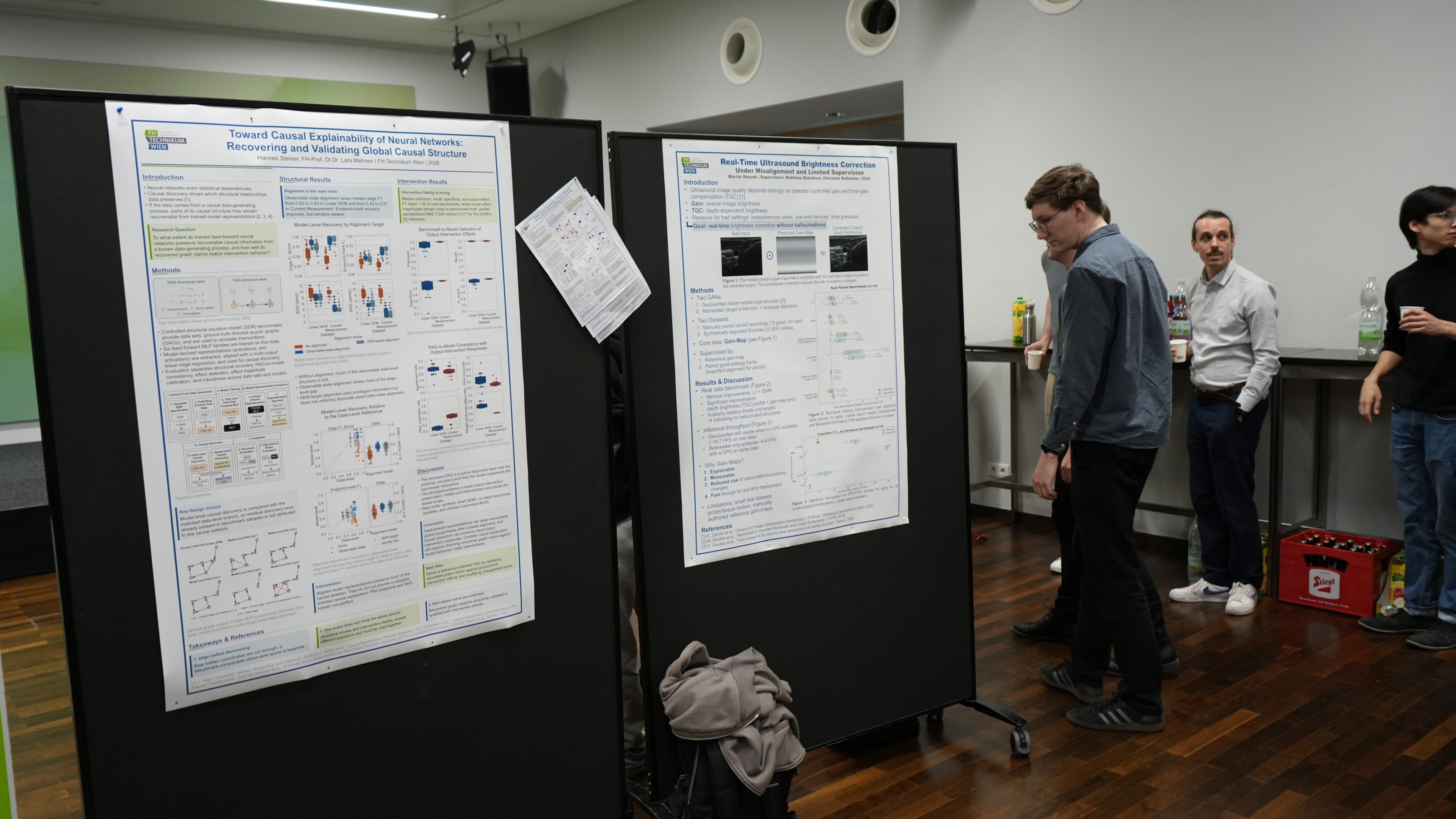 In einem Raum mit Holzboden stehen mehrere Personen vor zwei großen wissenschaftlichen Plakaten, die an schwarzen Tafeln angebracht sind. Auf dem Boden liegen Papiere und Taschen, und im Hintergrund sind auf einer Theke Getränke zu sehen.