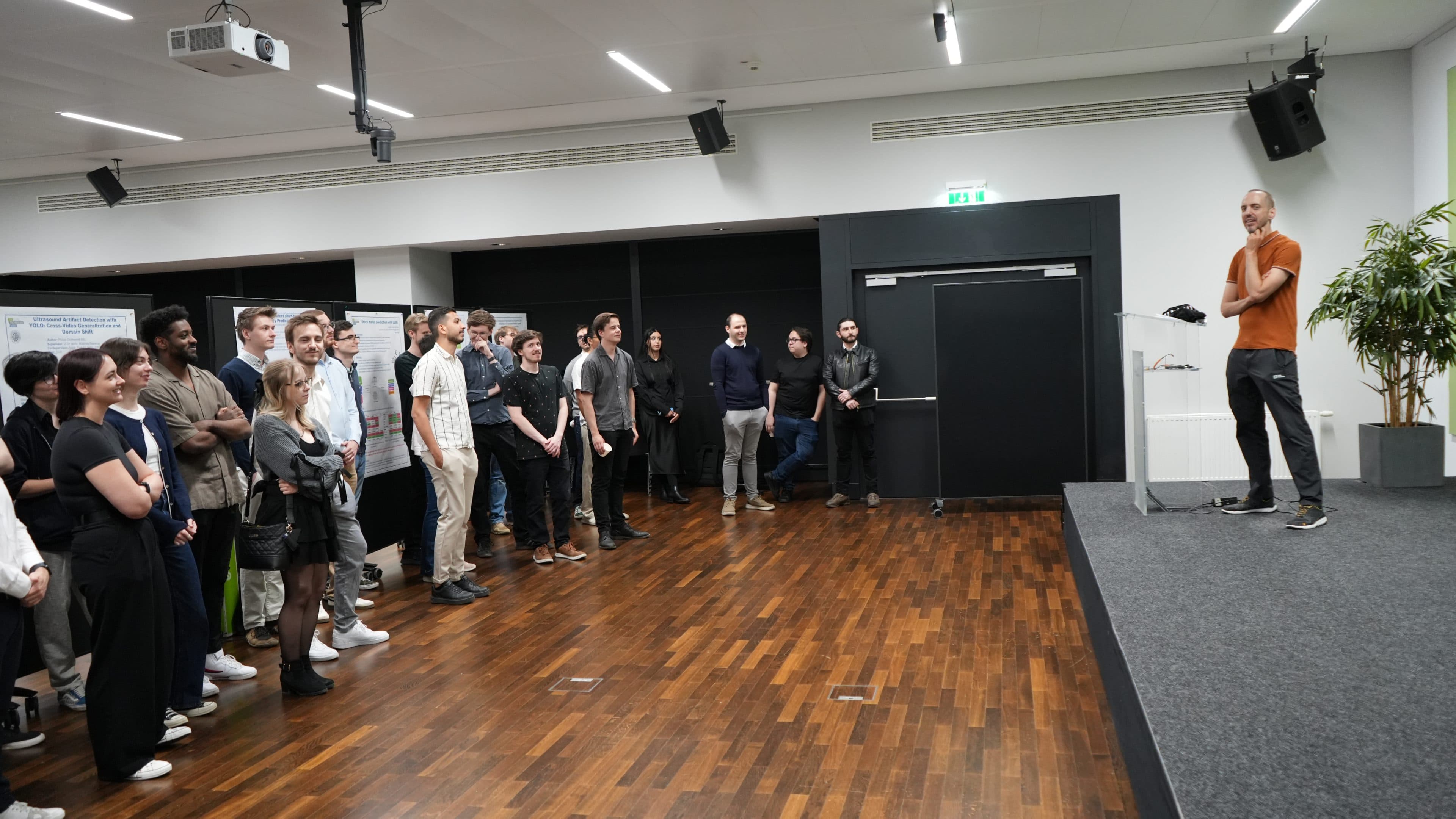 Ein Mann in einem orangefarbenen Hemd spricht von einem Podium auf einer Bühne zu einem aufmerksamen Publikum, das in einem modernen Raum mit Holzböden und weißen Wänden steht.