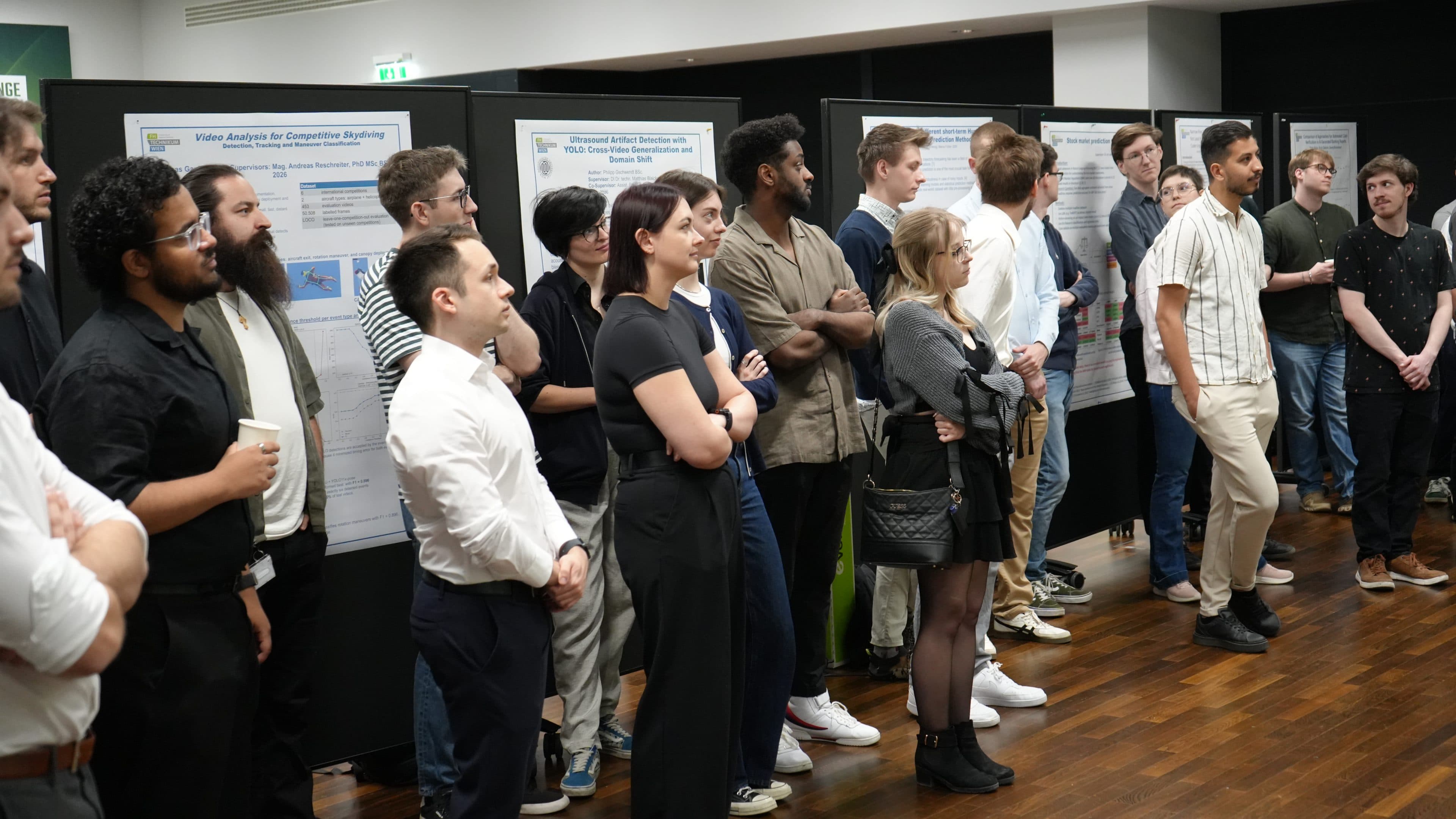Eine Gruppe von Menschen steht aufmerksam vor großen Ausstellungstafeln mit Forschungsplakaten in einem hell erleuchteten Innenraum, was auf eine fachliche oder akademische Veranstaltung hindeutet. Die meisten sind leger gekleidet und hören einem Redner zu.