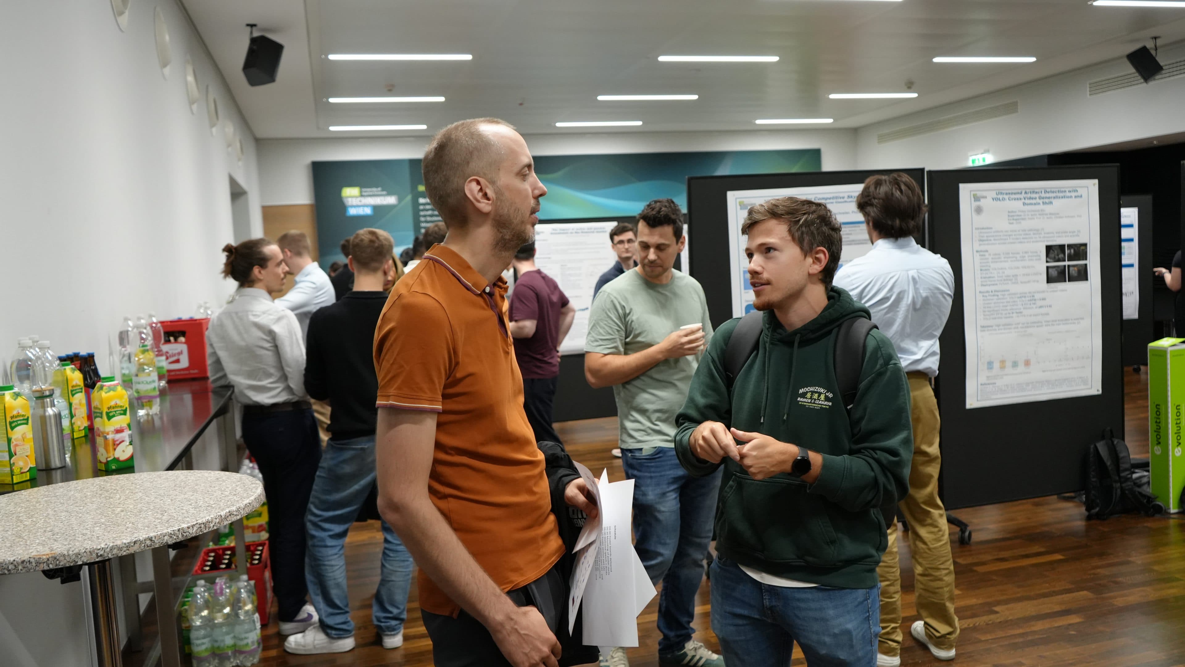 Die Teilnehmer einer Konferenz oder eines Seminars unterhalten sich in Gruppen in der Nähe von Plakaten und bei den Erfrischungen. Zwei Männer im Vordergrund sind in ein Gespräch vertieft, während andere im Hintergrund plaudern und sich die Plakate ansehen.