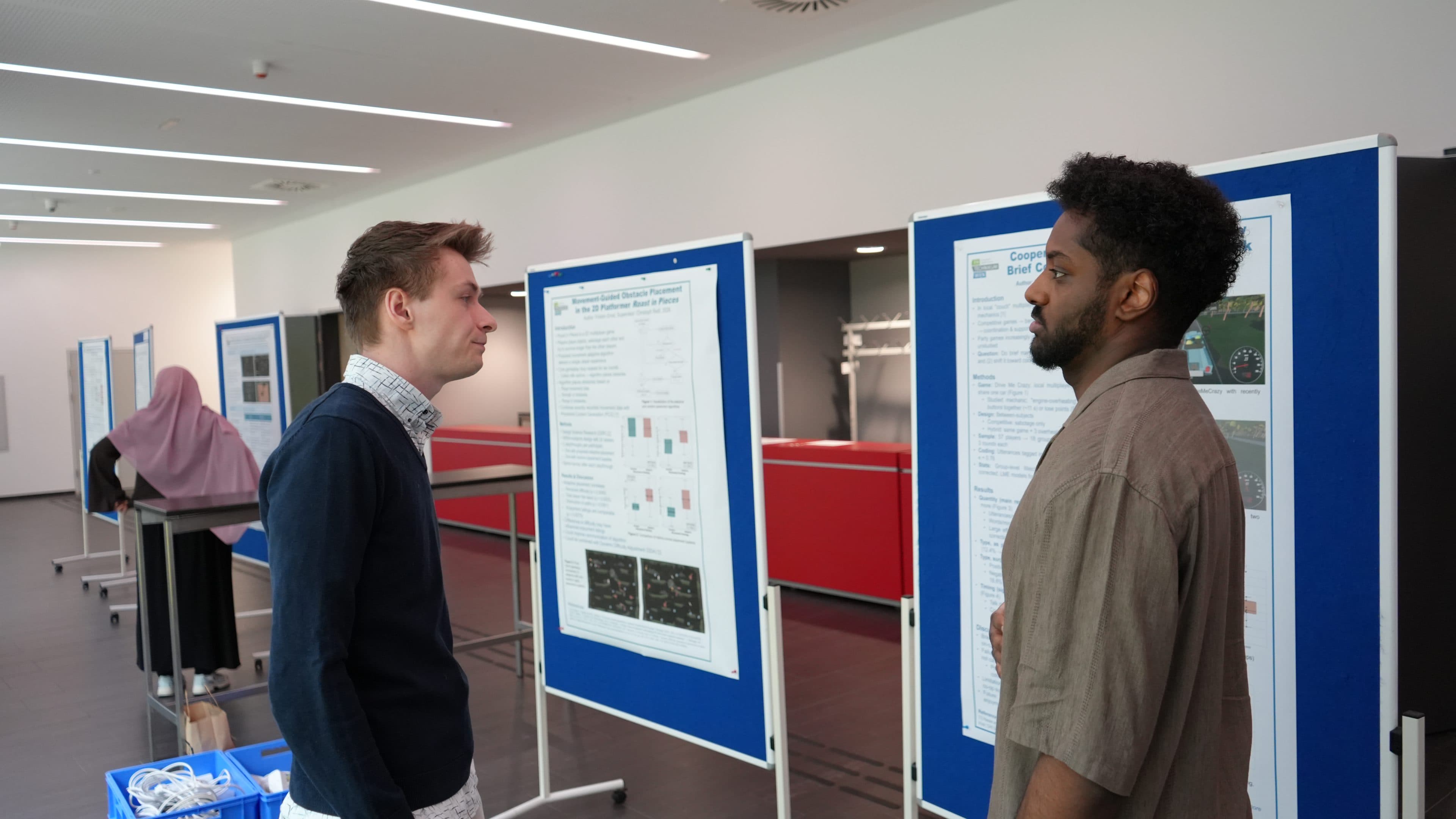 Zwei Männer stehen sich gegenüber und unterhalten sich vor blauen Ausstellungstafeln mit wissenschaftlichen Postern. Im Hintergrund liest eine Frau mit einem rosa Hijab ein weiteres Poster. Der Schauplatz scheint ein akademischer Raum oder ein Konferenzsaal zu sein.