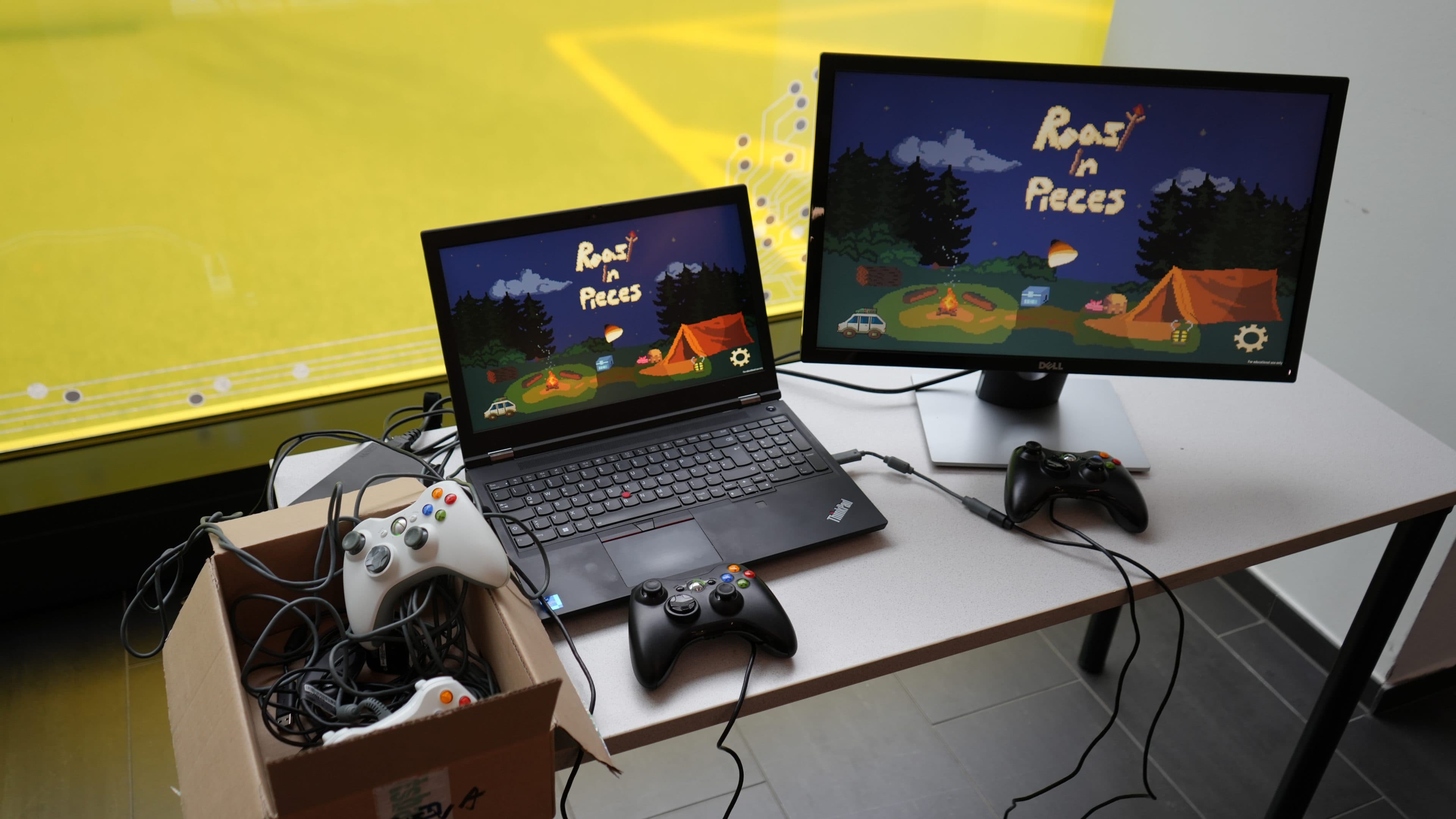 Auf einem Tisch stehen ein Laptop und ein Monitor, auf denen das Videospiel „Roast in Pieces“ läuft; daneben liegen zwei Spielcontroller und eine Schachtel mit weiteren Controllern und Kabeln. Die Aufstellung befindet sich in der Nähe eines Fensters mit Blick auf eine Grünfläche.