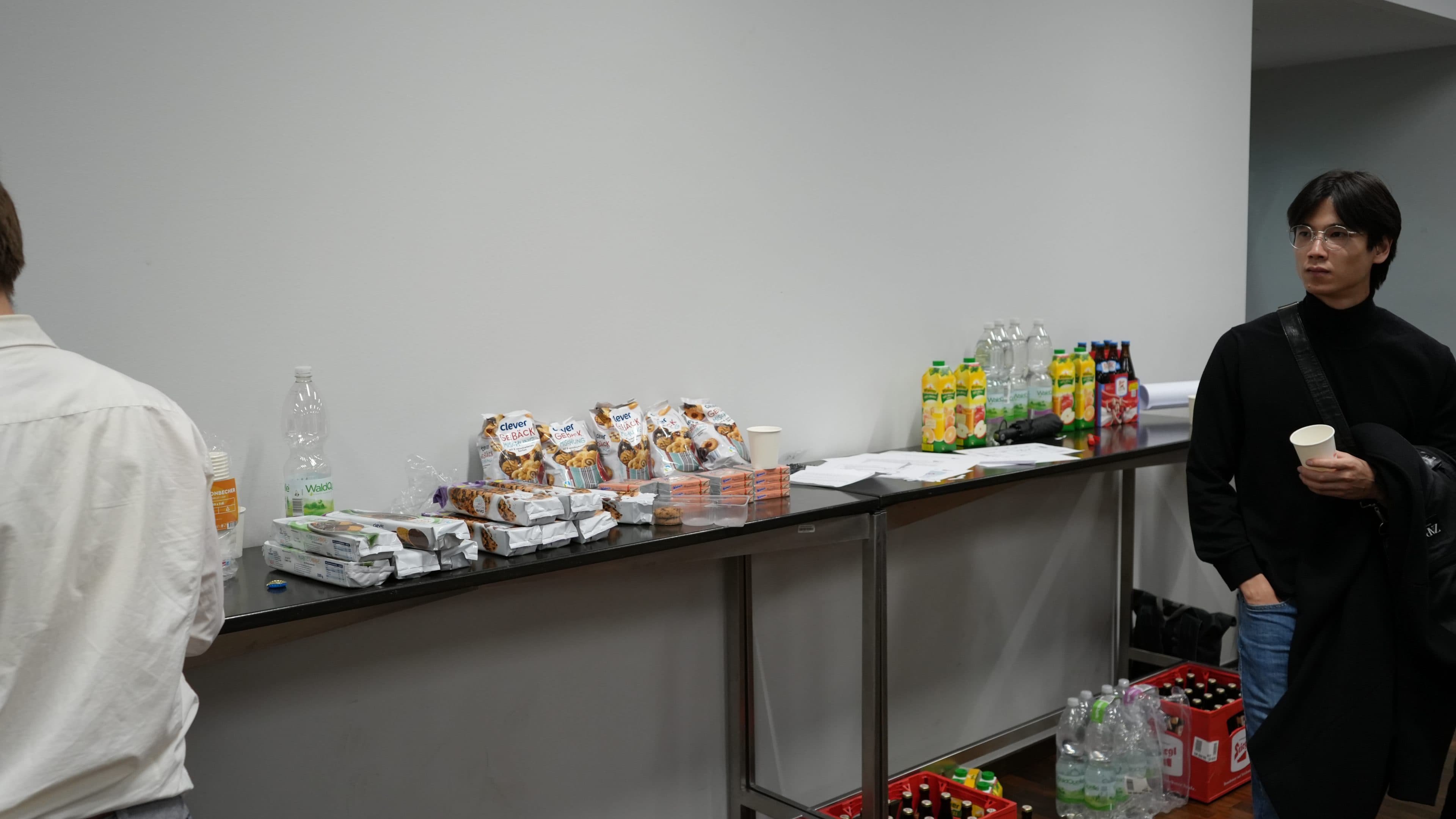 Ein Buffettisch, auf dem Snacks, Saftpäckchen, Wasserflaschen und Pappbecher aufgereiht sind. In der Nähe stehen mehrere Personen; eine Person in einer schwarzen Jacke hält einen Becher in der Hand und schaut nach links.