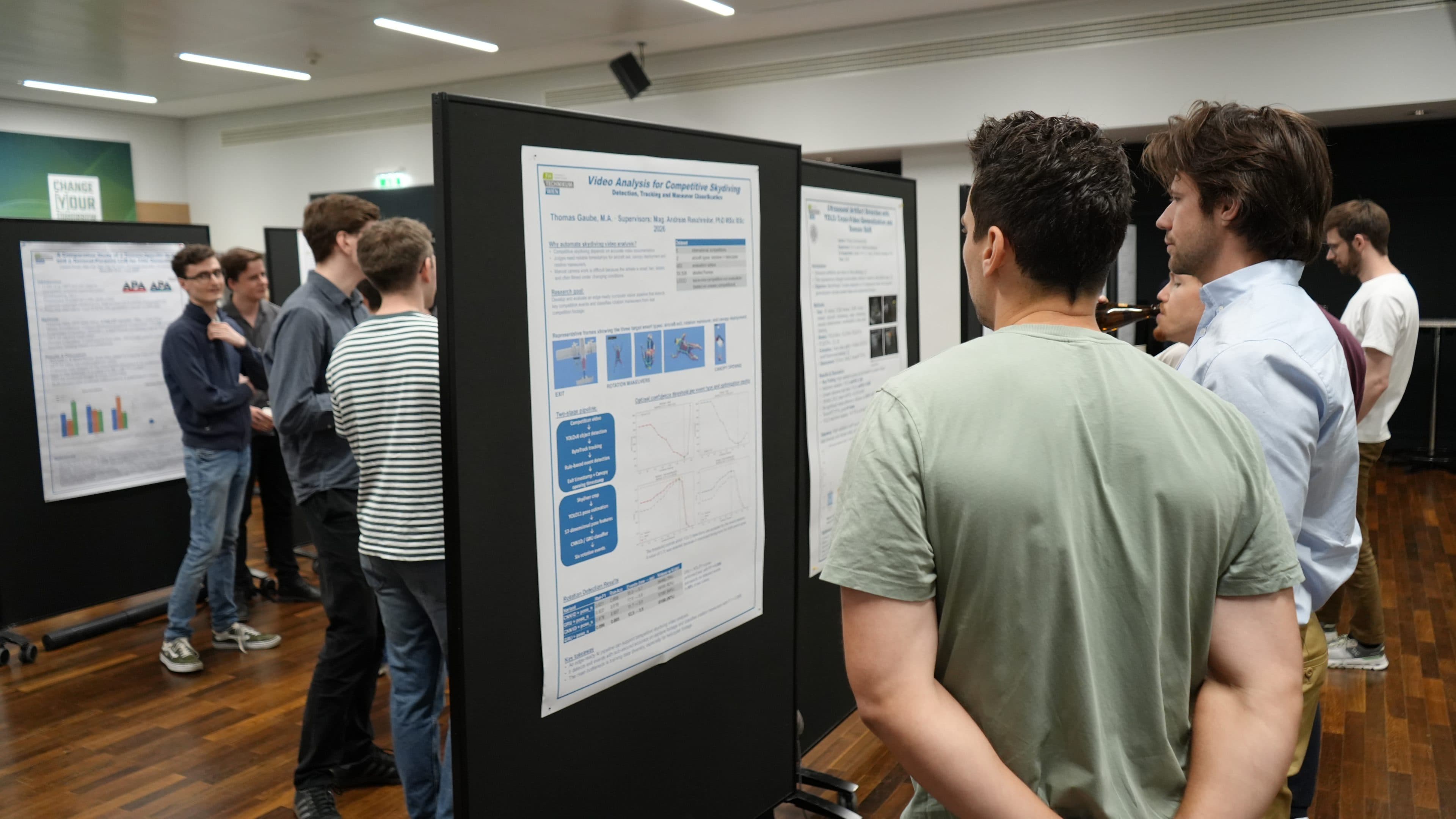 Eine Gruppe von Menschen steht in einem Raum und betrachtet und bespricht wissenschaftliche Poster, die bei einer akademischen oder wissenschaftlichen Veranstaltung an schwarzen Tafeln ausgestellt sind.
