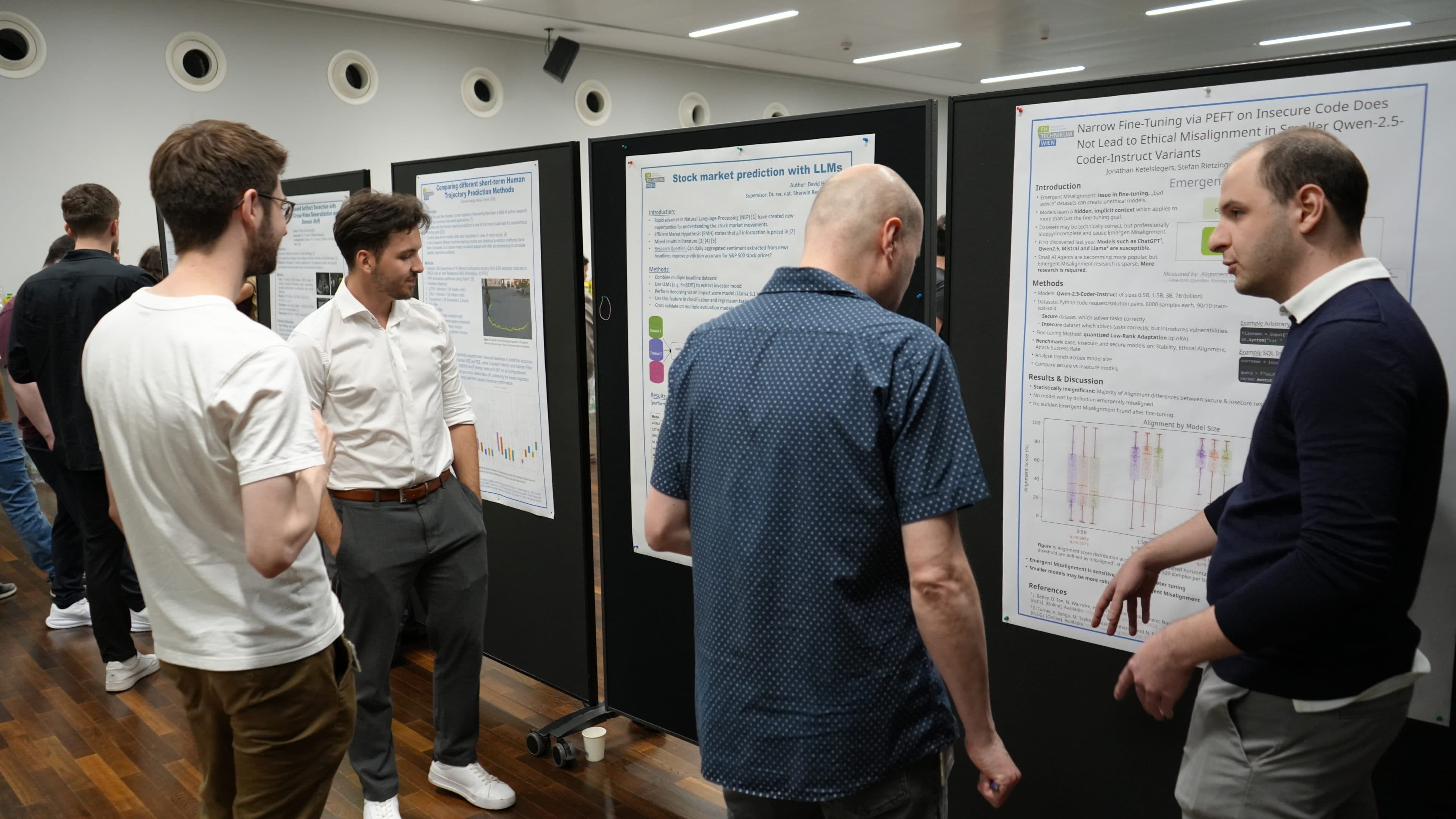 Vier Männer stehen bei einer wissenschaftlichen Konferenz oder Veranstaltung und besprechen Forschungsplakate. Die Plakate sind an schwarzen Ausstellungstafeln in einem hell erleuchteten Raum mit Holzboden und weißen Wänden angebracht.