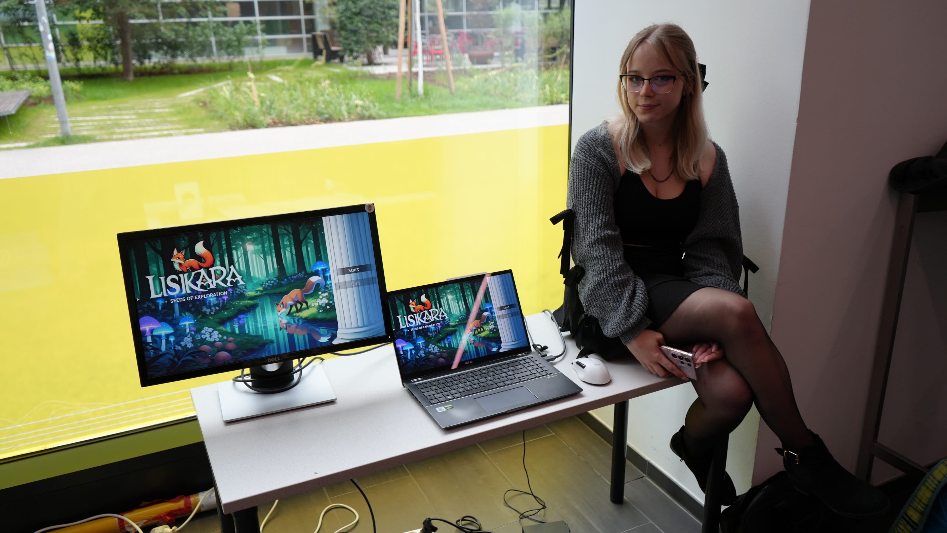 Eine Frau mit blonden Haaren und Brille sitzt an einem weißen Schreibtisch neben einem Monitor und einem Laptop, auf denen beide ein farbenfrohes Waldbild mit dem Titel „LISIKARA“ zu sehen ist. Durch ein großes Fenster ist hinter ihr eine grüne Außenanlage zu sehen.