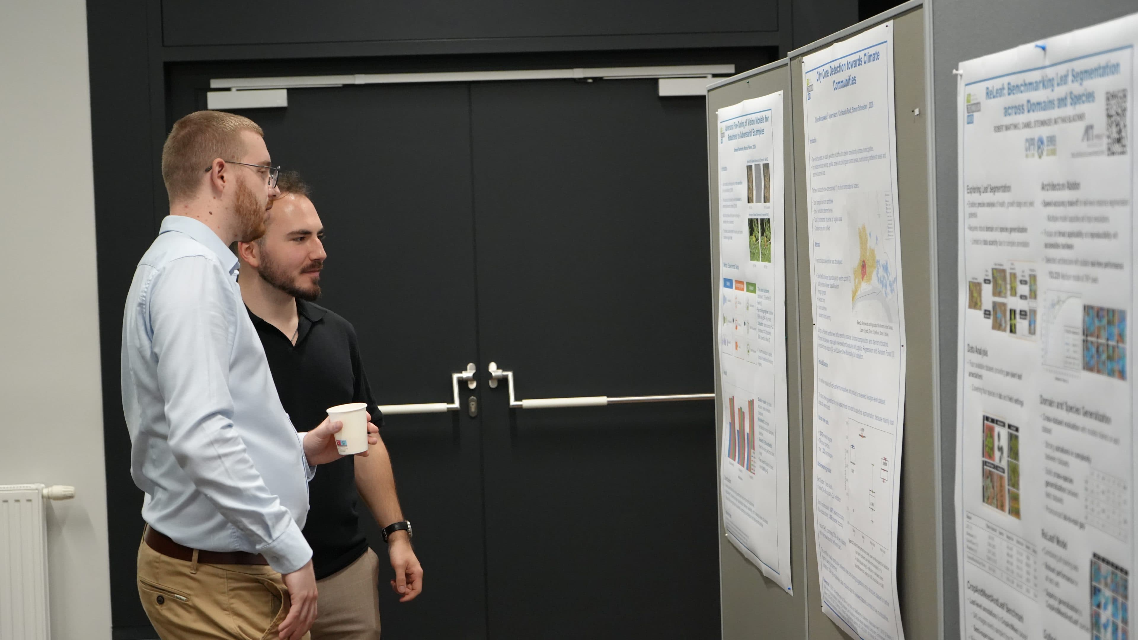 Zwei Männer stehen vor wissenschaftlichen Postern an Ausstellungstafeln und besprechen deren Inhalt. Einer hält eine Tasse in der Hand. Beide wirken konzentriert, im Hintergrund sind geschlossene Flügeltüren und eine graue Wand zu sehen.
