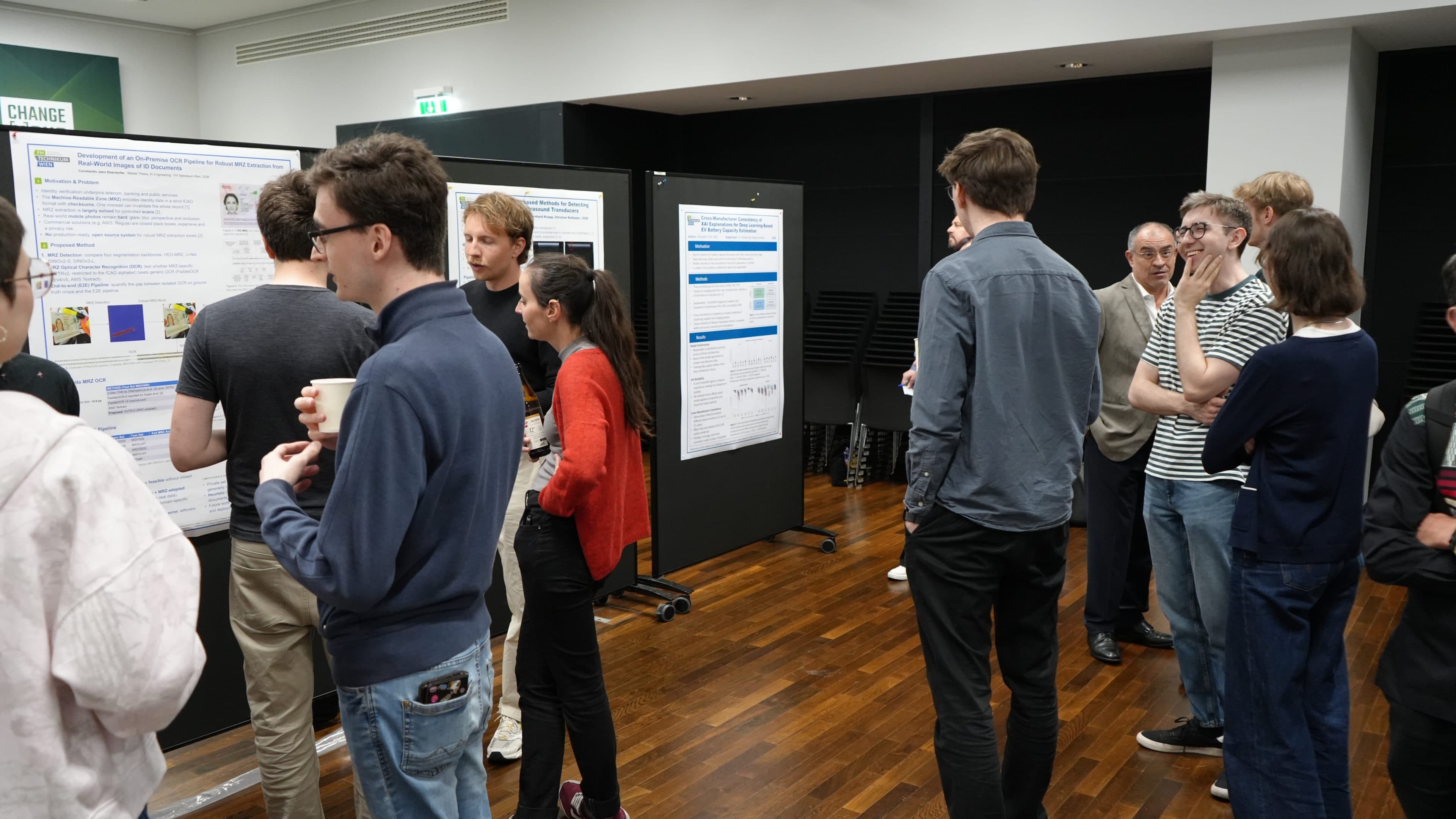 Eine Gruppe von Menschen steht in einem Raum, betrachtet Forschungsplakate, die an schwarzen Tafeln angebracht sind, und diskutiert darüber; einige unterhalten sich zu zweit oder in kleinen Gruppen auf einem Holzboden.