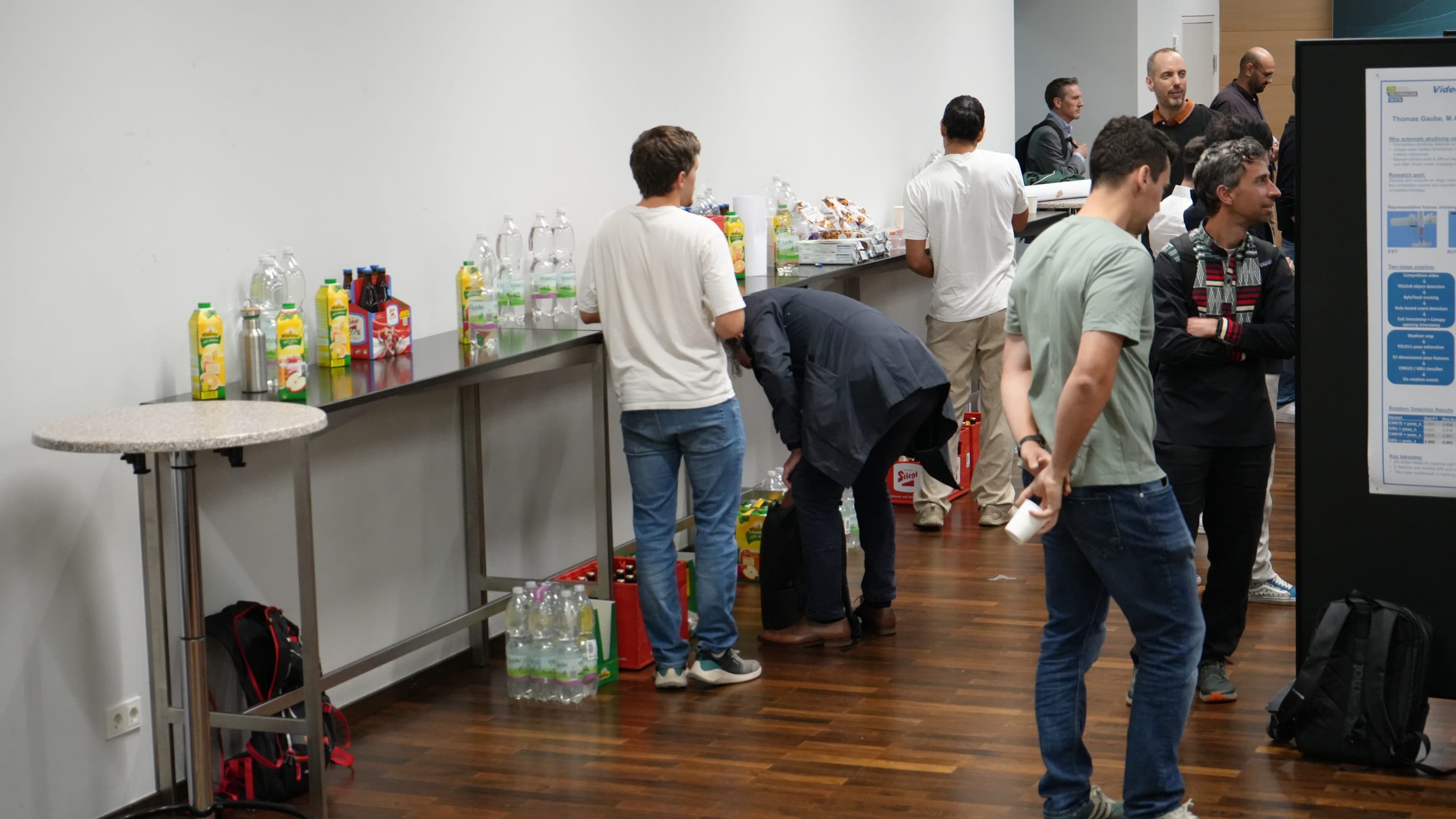 Bei einer Veranstaltung in einem Raum mit Holzboden versammeln sich die Leute um einen Tisch mit Erfrischungen, an dem Saftflaschen, Wasser und Snacks bereitstehen. Einige bedienen sich selbst, während andere sich unterhalten oder daneben stehen.
